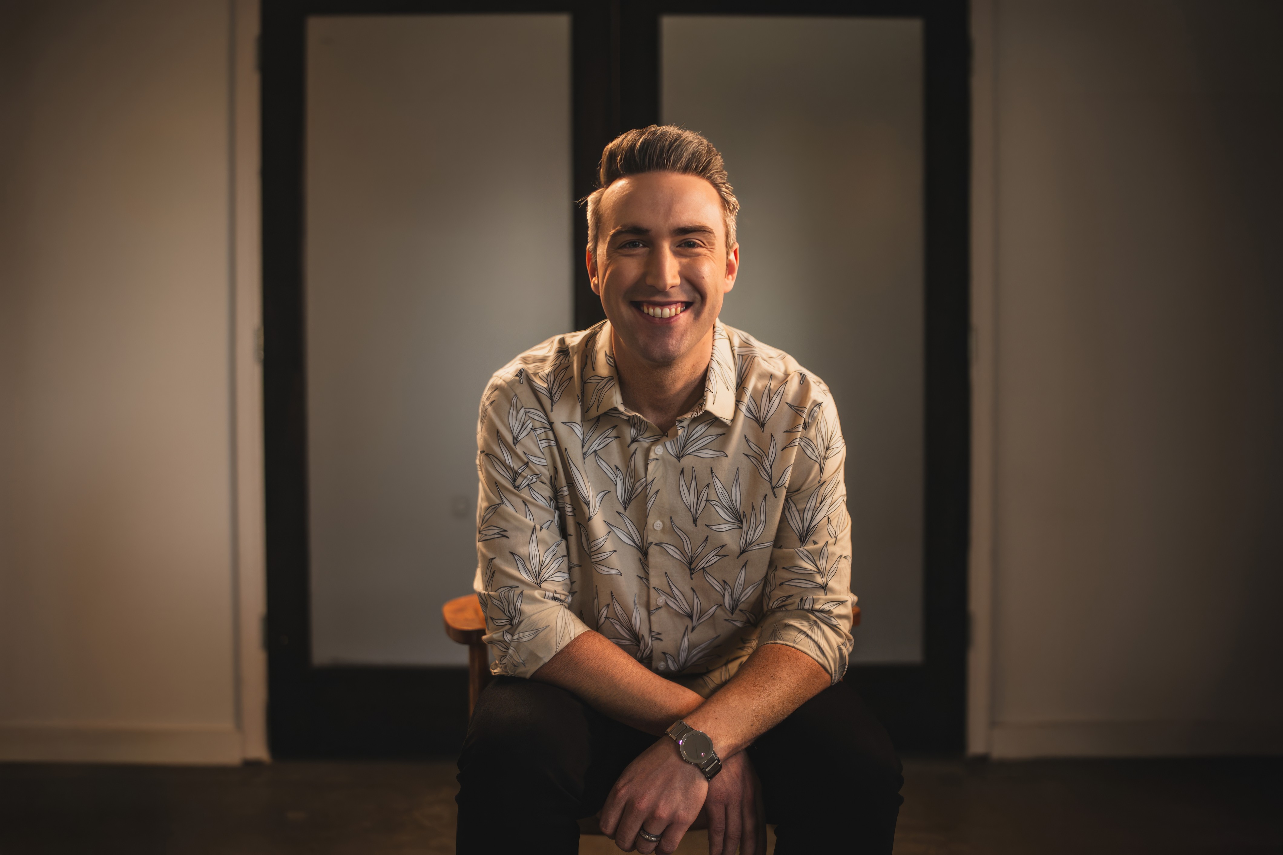 Smiling man with short hair and patterned shirt sitting in front of a dark door, arms crossed, in a warmly lit room.