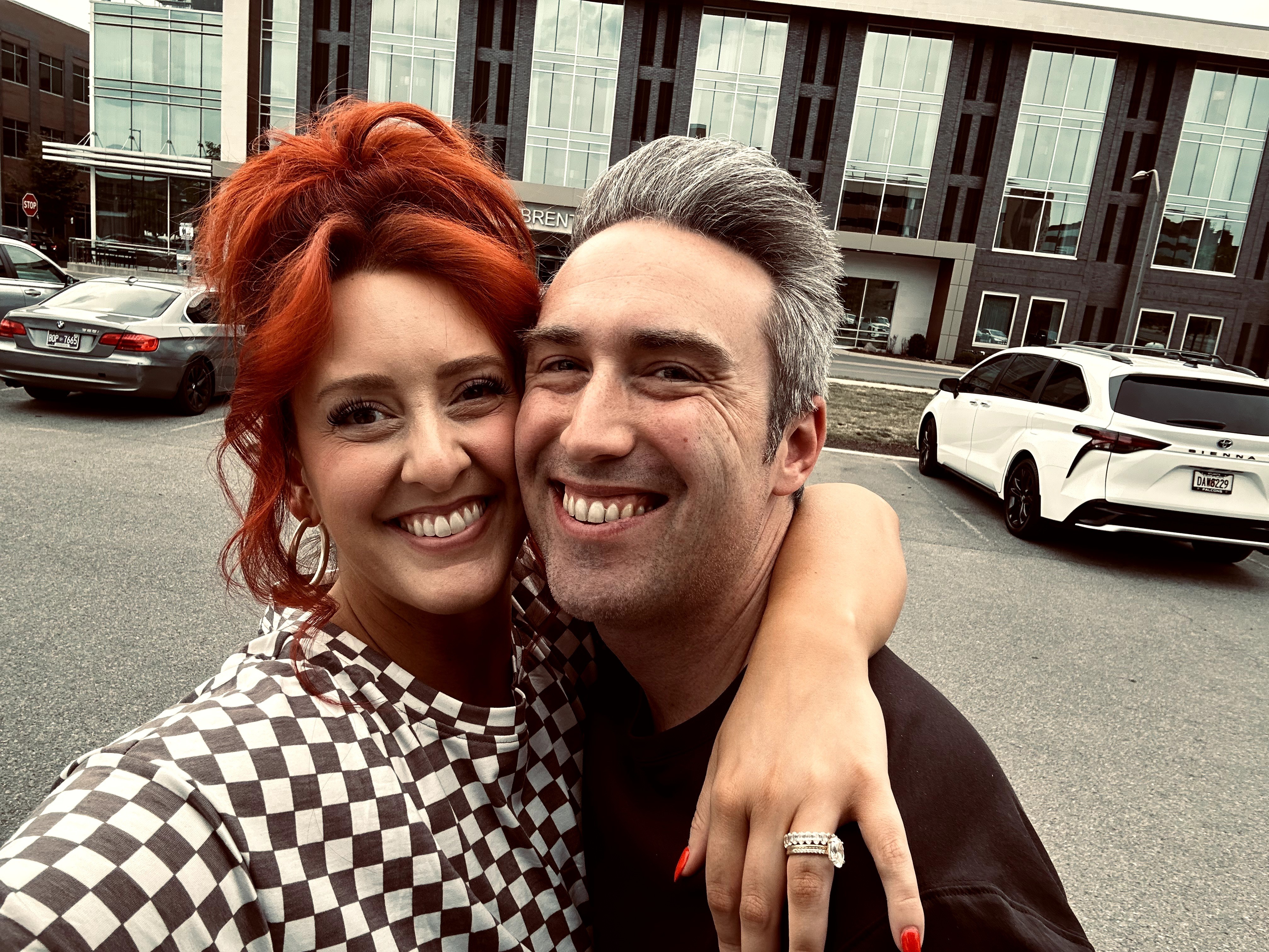 Two people smiling and hugging outdoors in front of modern buildings and parked cars.