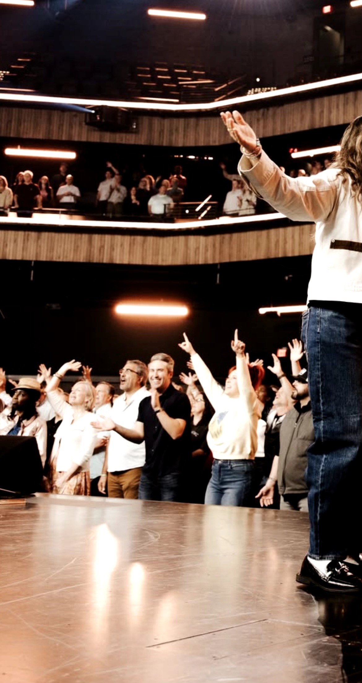 Person on stage gesturing to audience in a large indoor venue with multiple levels and audience members raising hands.