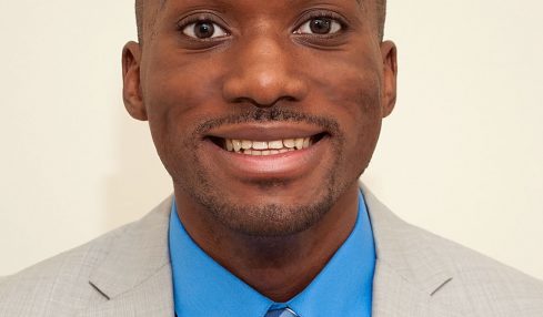 Portrait of a man in a light gray suit, blue shirt, and striped tie, smiling against a plain background.