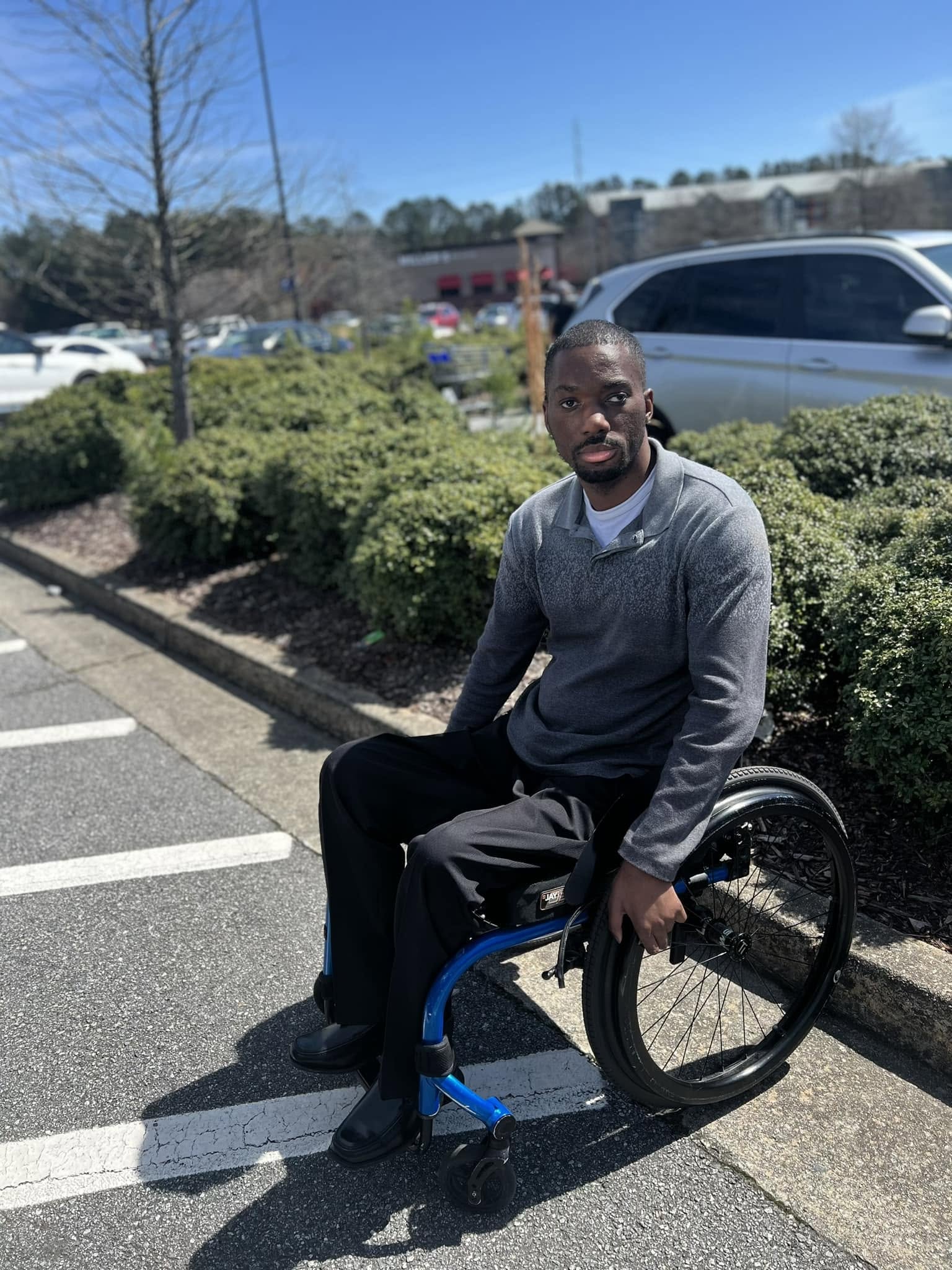 Man sitting in a wheelchair outdoors in a parking lot with bushes and cars in the background.