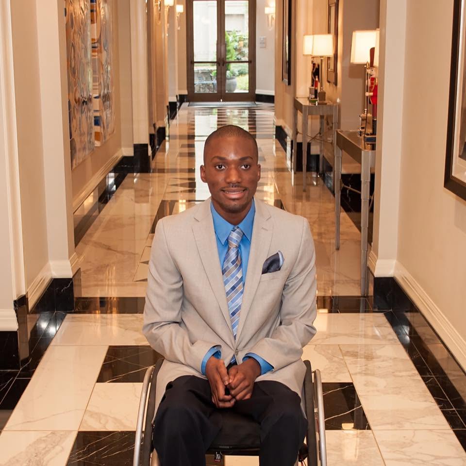 Young man in a wheelchair wearing a light gray suit, blue shirt, and striped tie, sitting in a hotel corridor.
