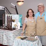 Two people stand behind a table with jewelry displays, smiling indoors. The woman has glasses and curly hair; the man wears a cap.