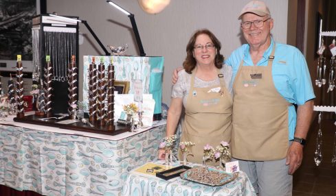 Two people stand behind a table with jewelry displays, smiling indoors. The woman has glasses and curly hair; the man wears a cap.