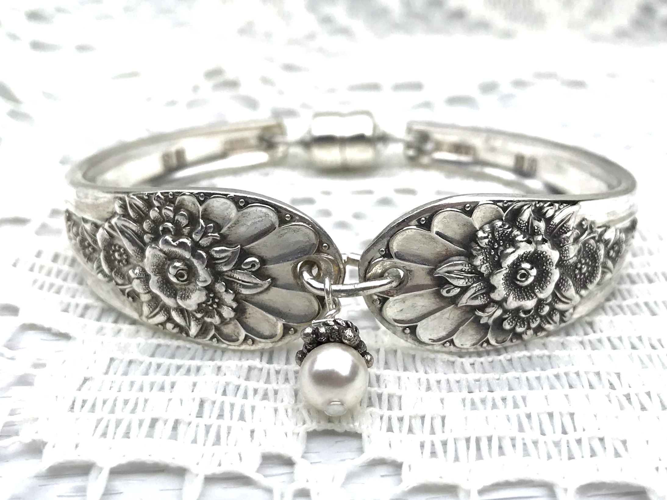 Silver bracelet with floral engravings and a dangling pearl charm, resting on textured fabric.