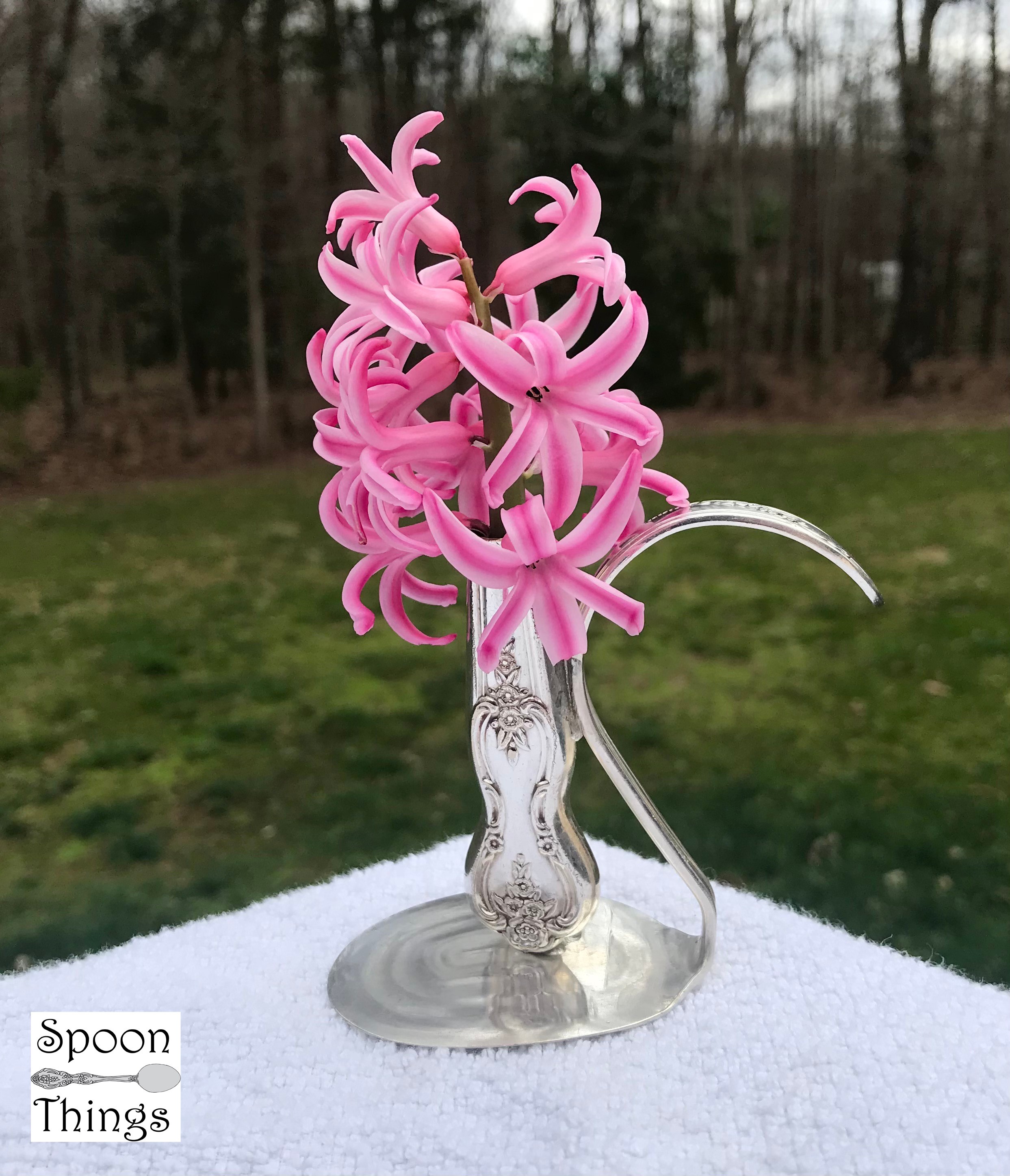 Pink curly glass sculpture on a silver stand outdoors with trees and grass in background.