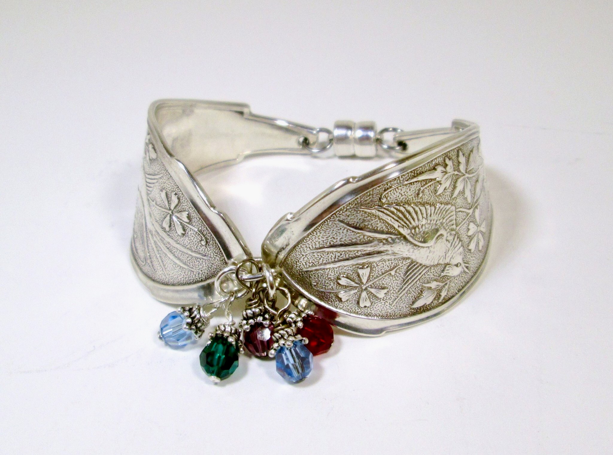 Silver bracelet with engraved floral patterns and dangling colorful gemstones, on a white background.