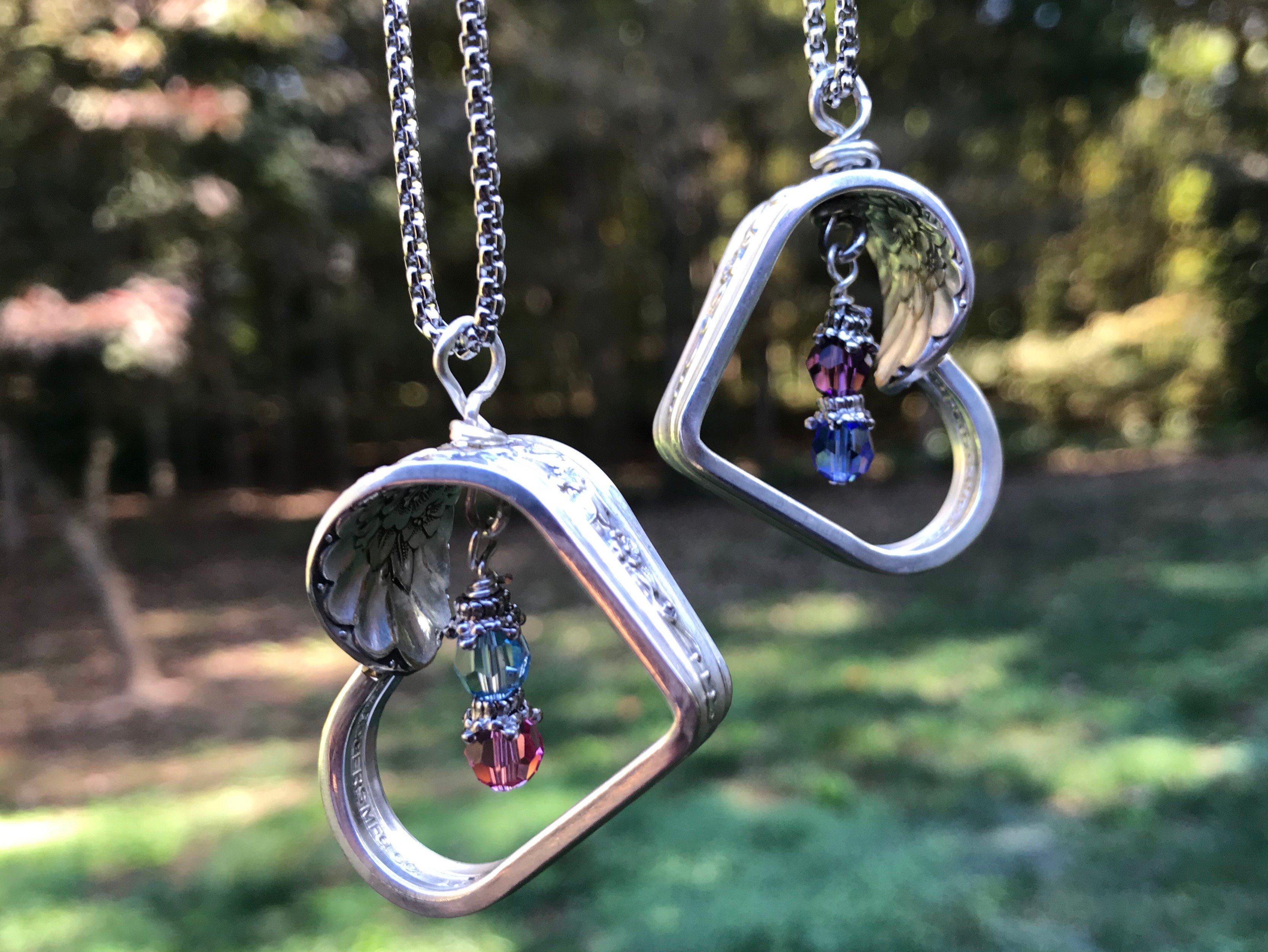 Two heart-shaped necklaces with beads hang outdoors against a blurred background of trees and grass.