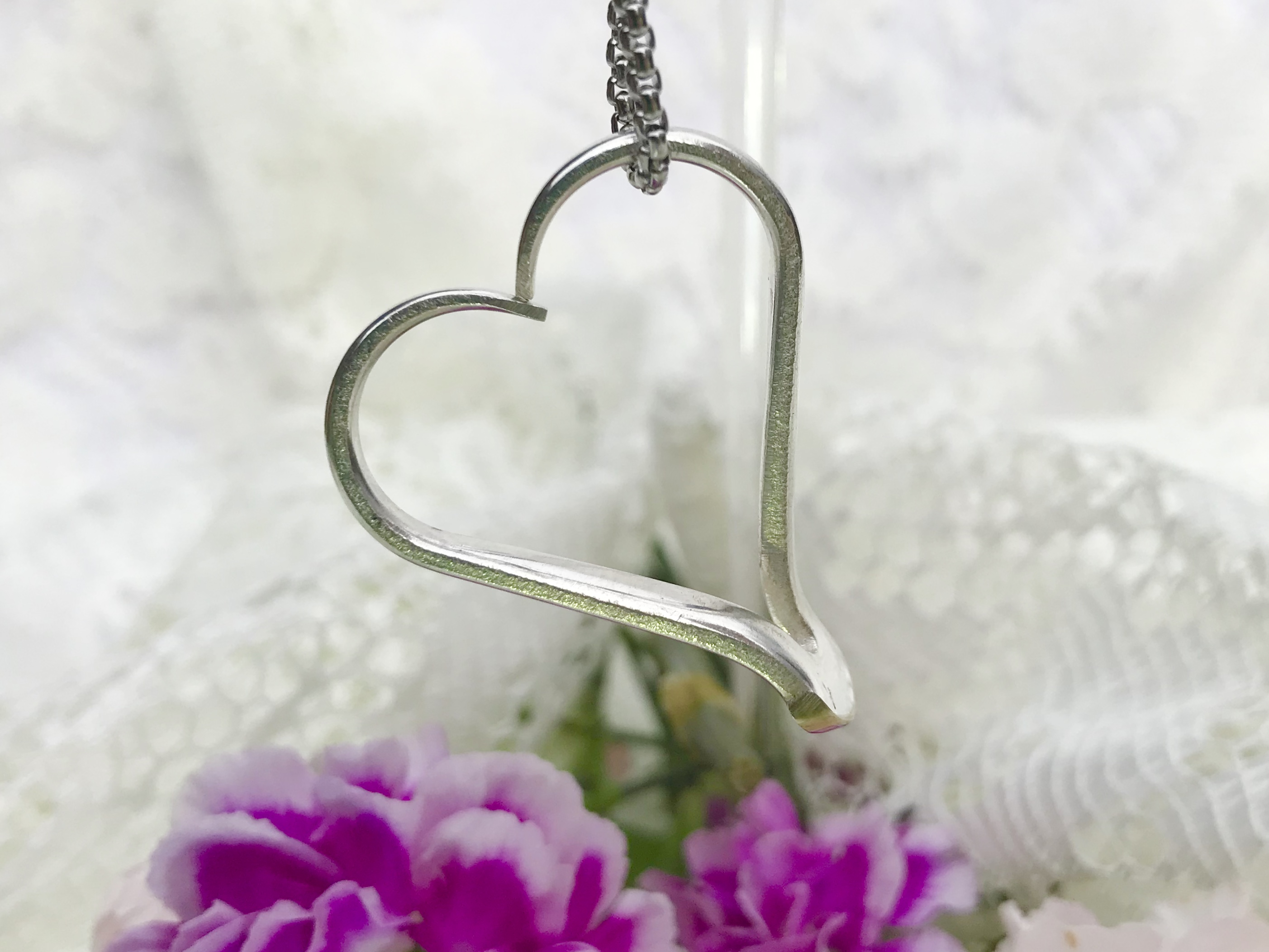 Heart-shaped metal necklace hanging against a light background with purple flowers nearby.