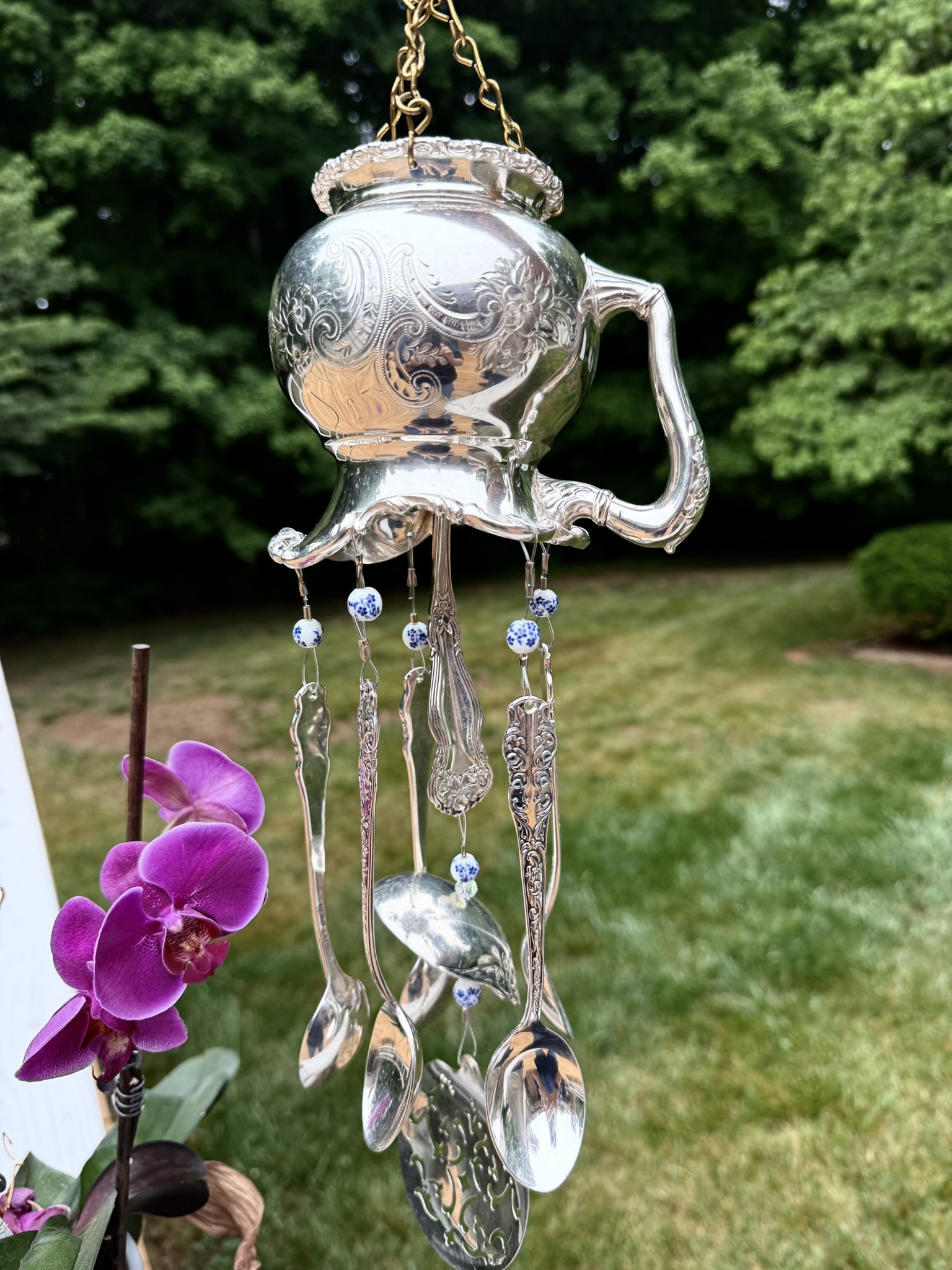 Teapot-shaped wind chime with hanging glass and metal pieces, outdoors with green trees and grass in background.