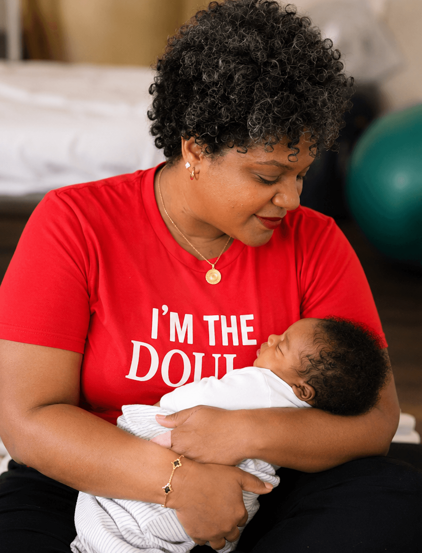 Woman holding a baby, wearing a red shirt with white text, in a room with a bed and exercise ball in background.