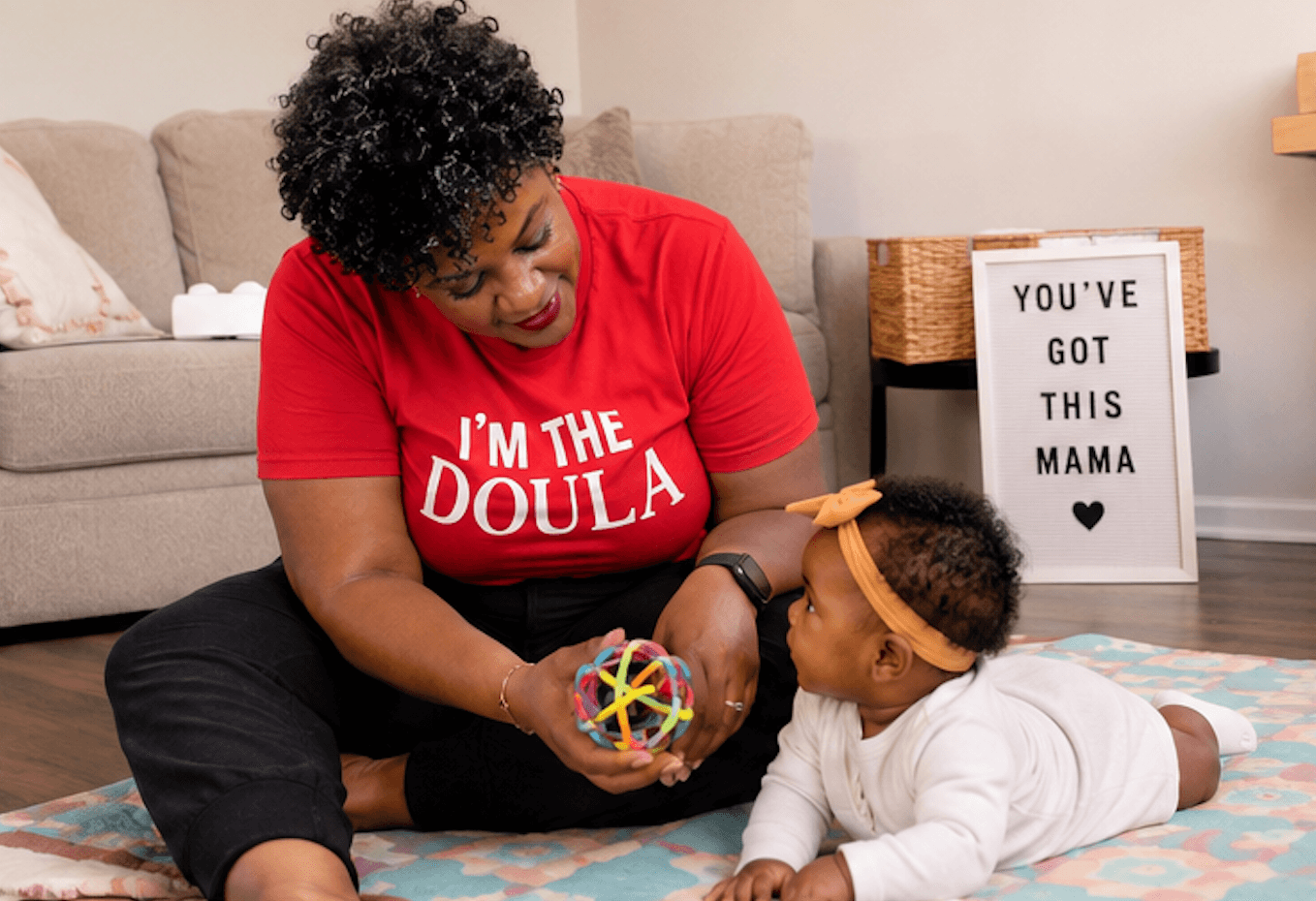 Adult woman and child playing with a colorful ball on a rug in a living room, with a sign reading 'You've got this mama'.