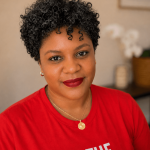 Woman with short curly hair wearing a red shirt with white text, in a room with a blurred background.