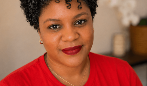 Woman with short curly hair wearing a red shirt with white text, in a room with a blurred background.