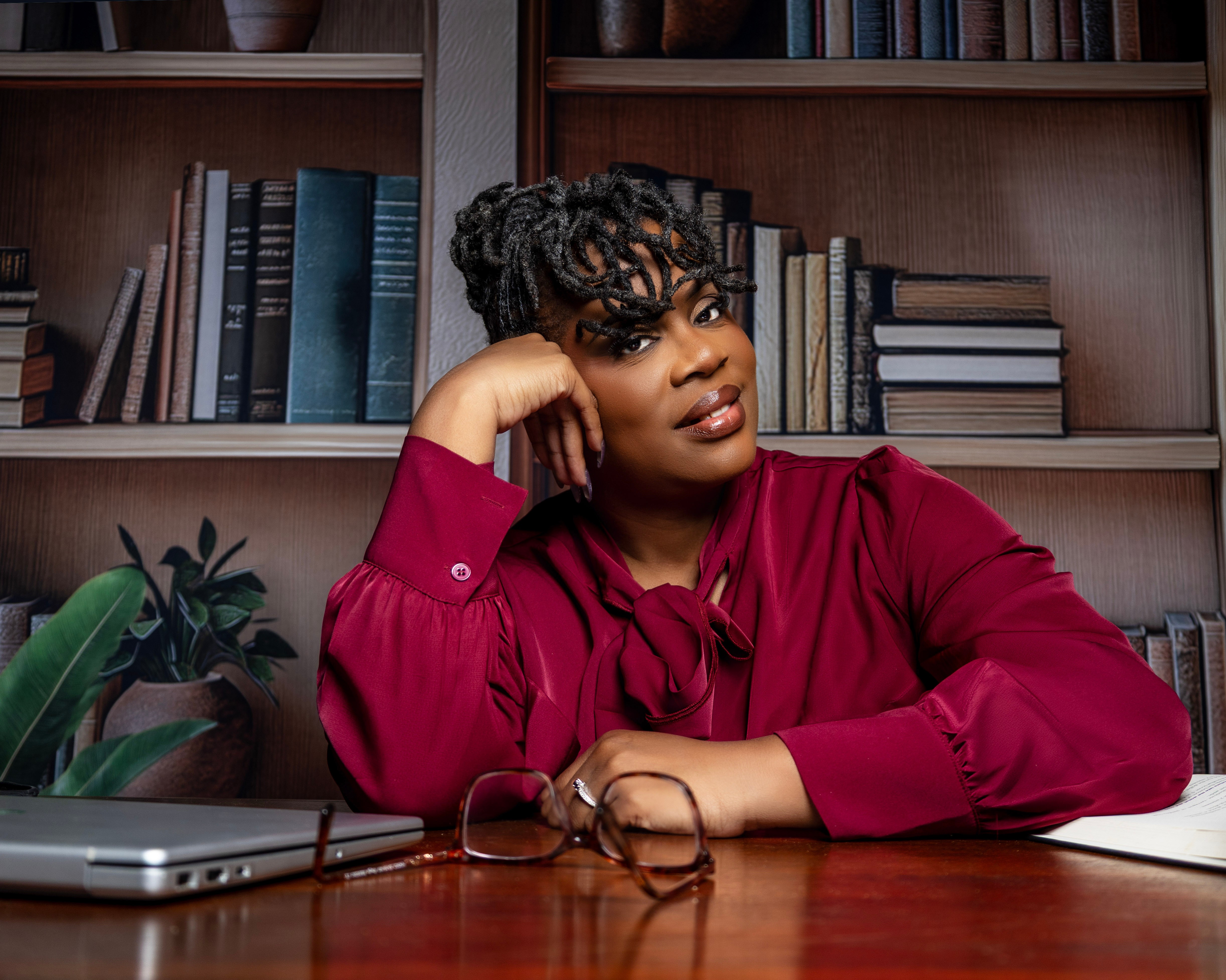Woman with curly hair in a red blouse sitting at a desk with bookshelves behind her.