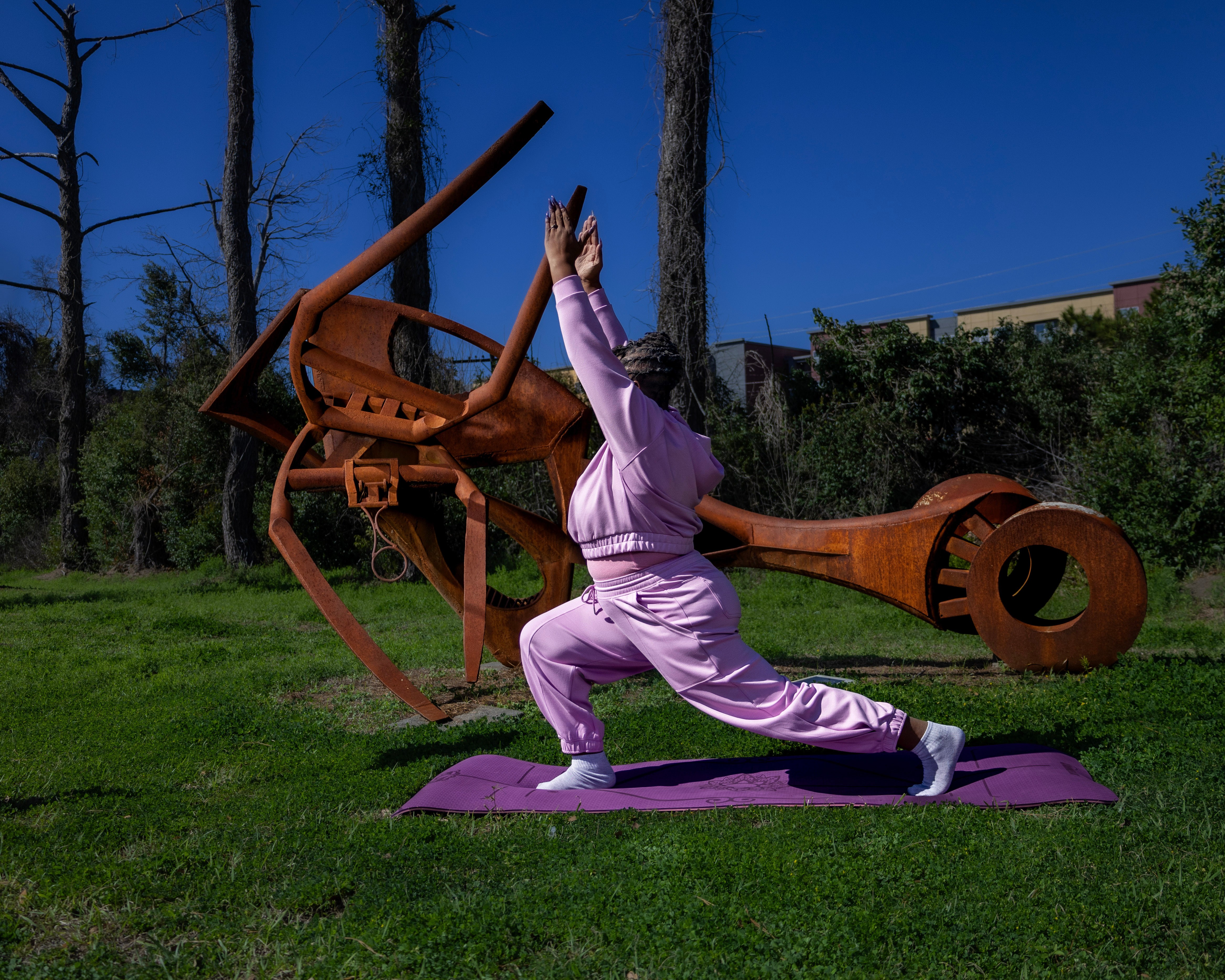 Person practicing yoga outdoors on a purple mat, with a large rust-colored sculpture and trees in the background.
