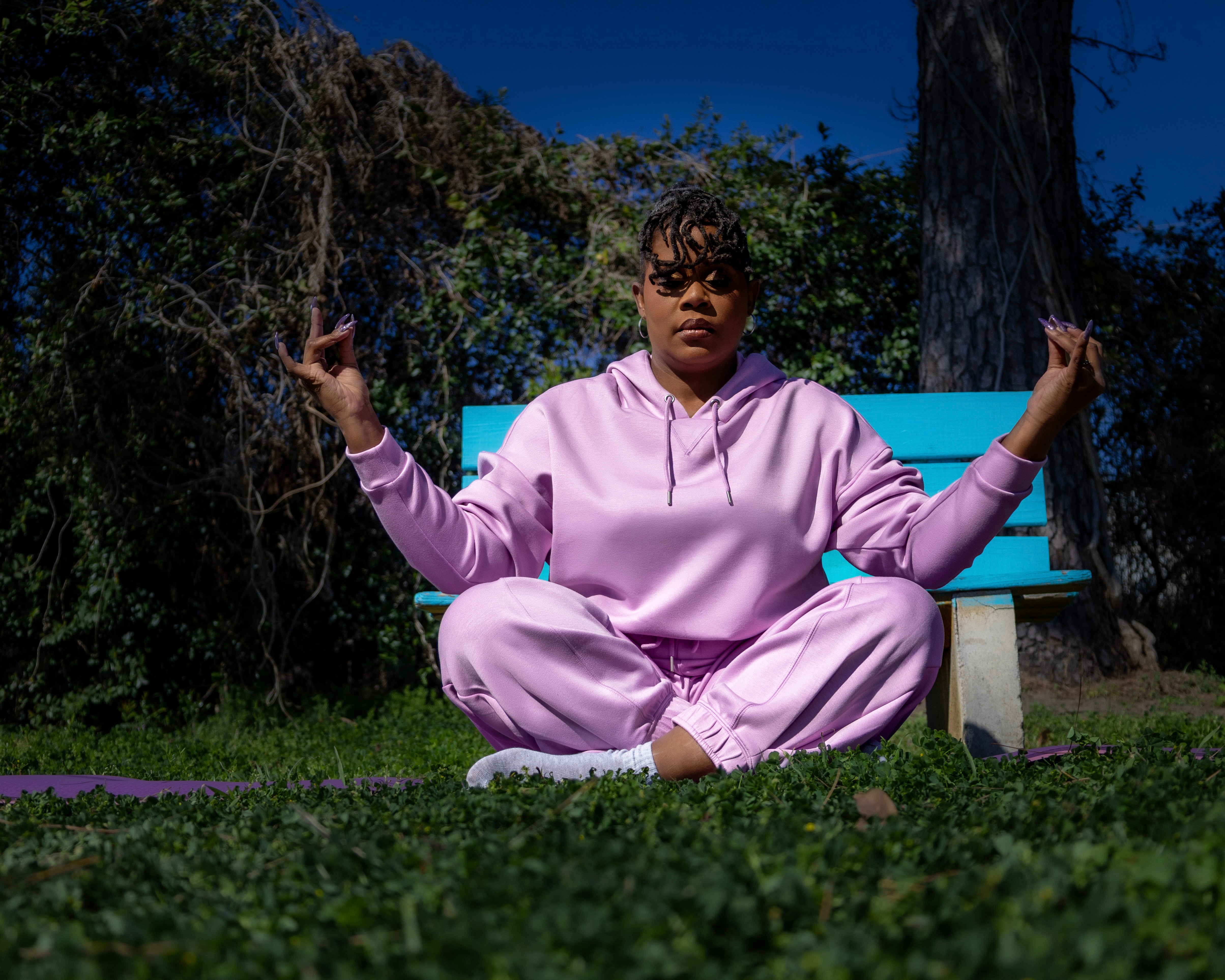 Person sitting cross-legged on grass, wearing pink hoodie and pants, holding tree branches, outdoors during daytime.