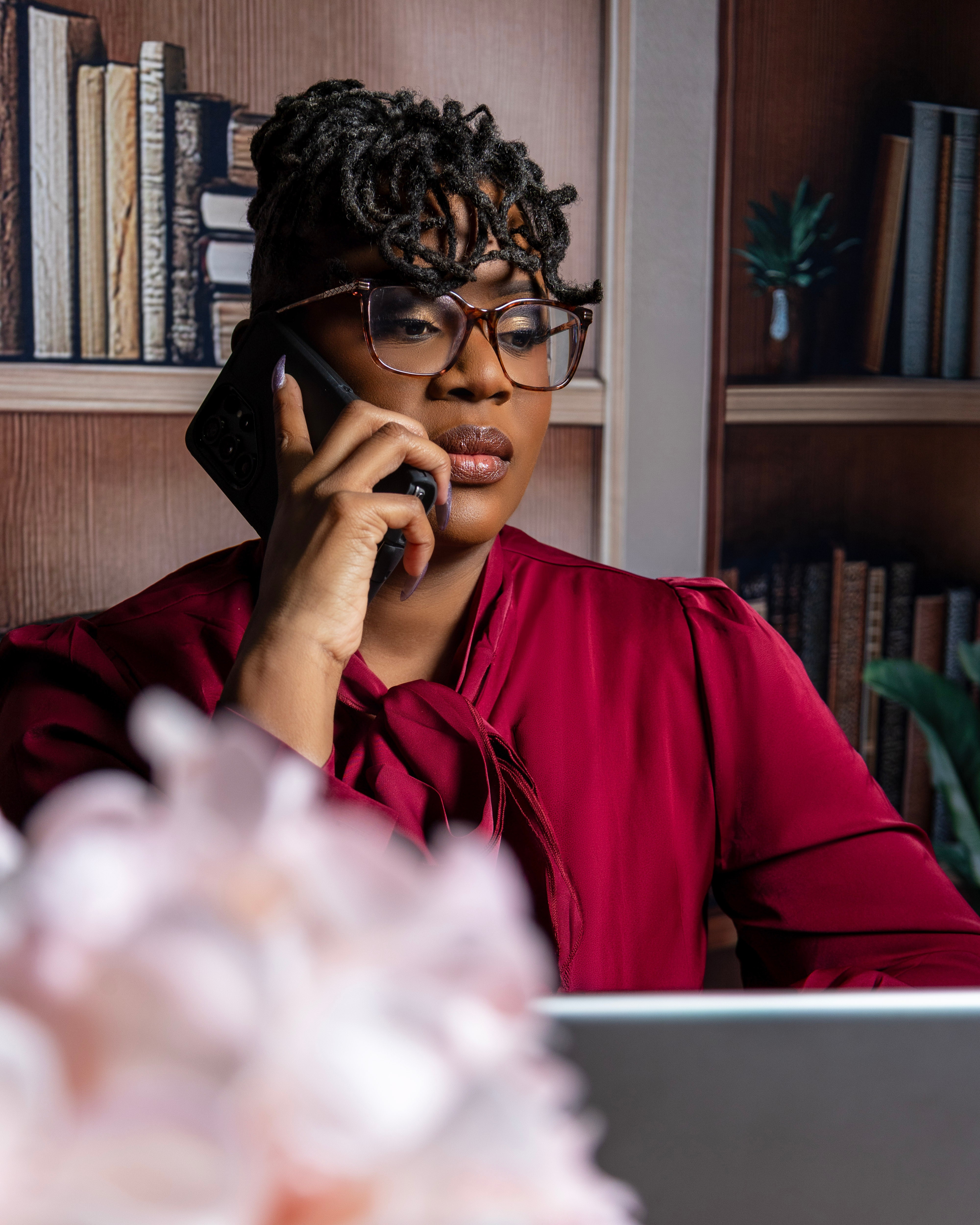 Woman with glasses and curly hair talking on phone in a library, wearing a red blouse, with bookshelves behind her.