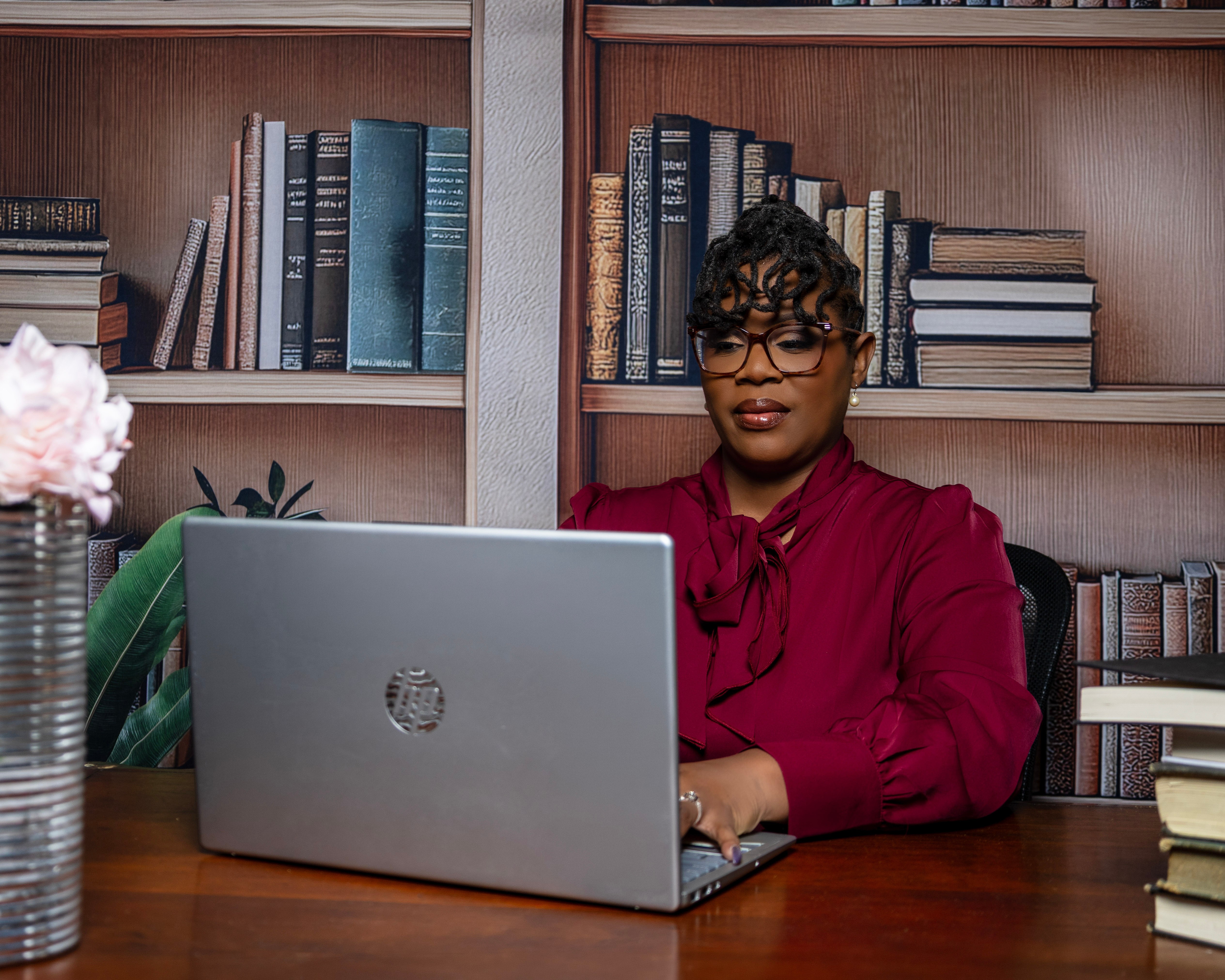 Woman in red blouse working on laptop at desk with bookshelves in background.
