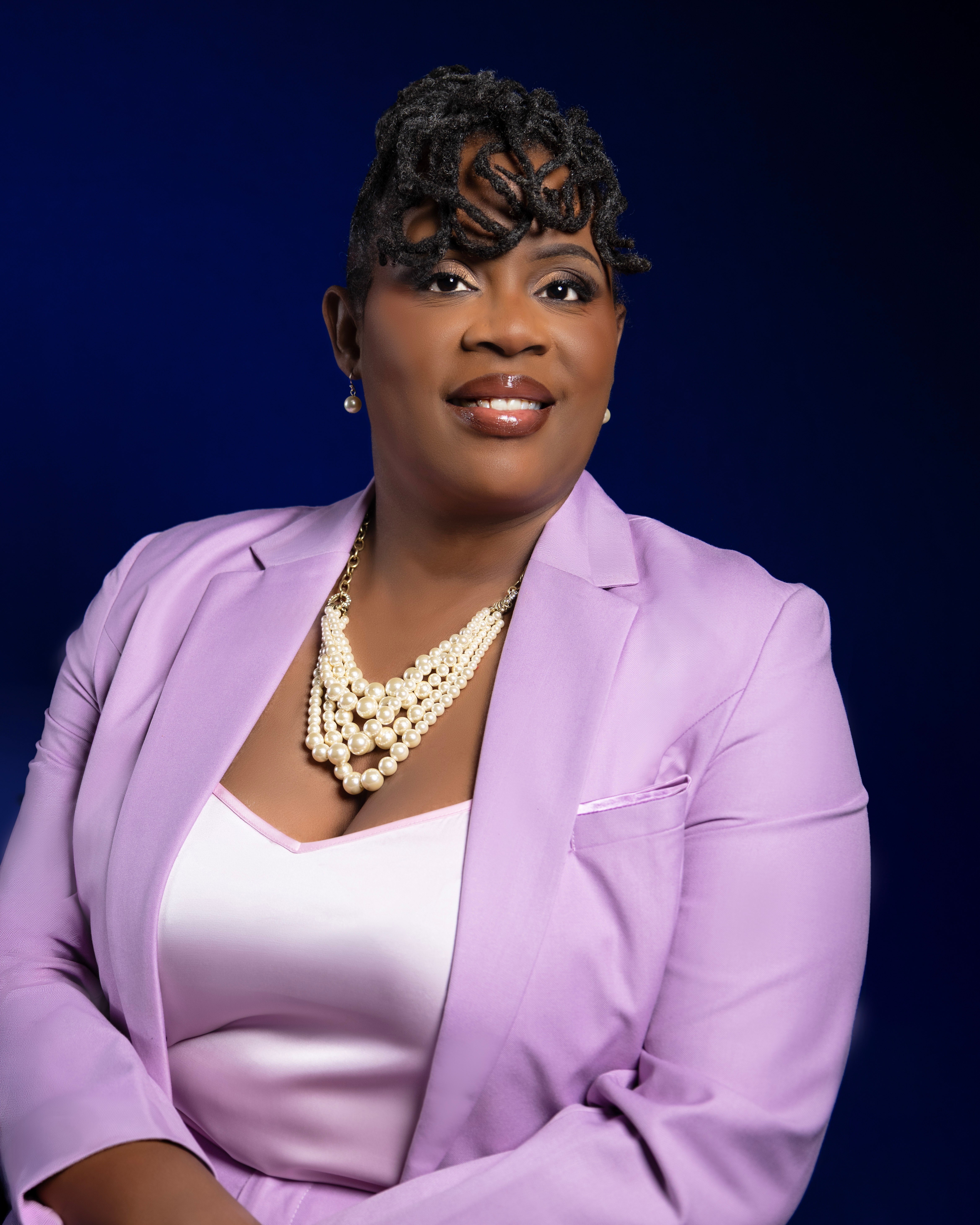 Portrait of a woman wearing a lavender blazer and pearl necklace, smiling against a dark background.
