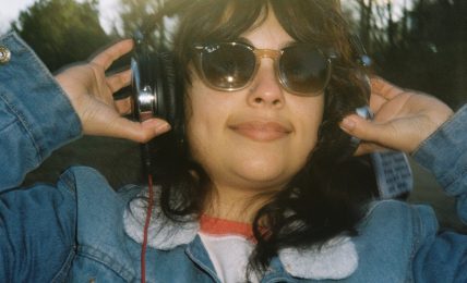 Person wearing sunglasses and headphones outdoors, smiling, with trees and sky in background.