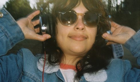 Person wearing sunglasses and headphones outdoors, smiling, with trees and sky in background.