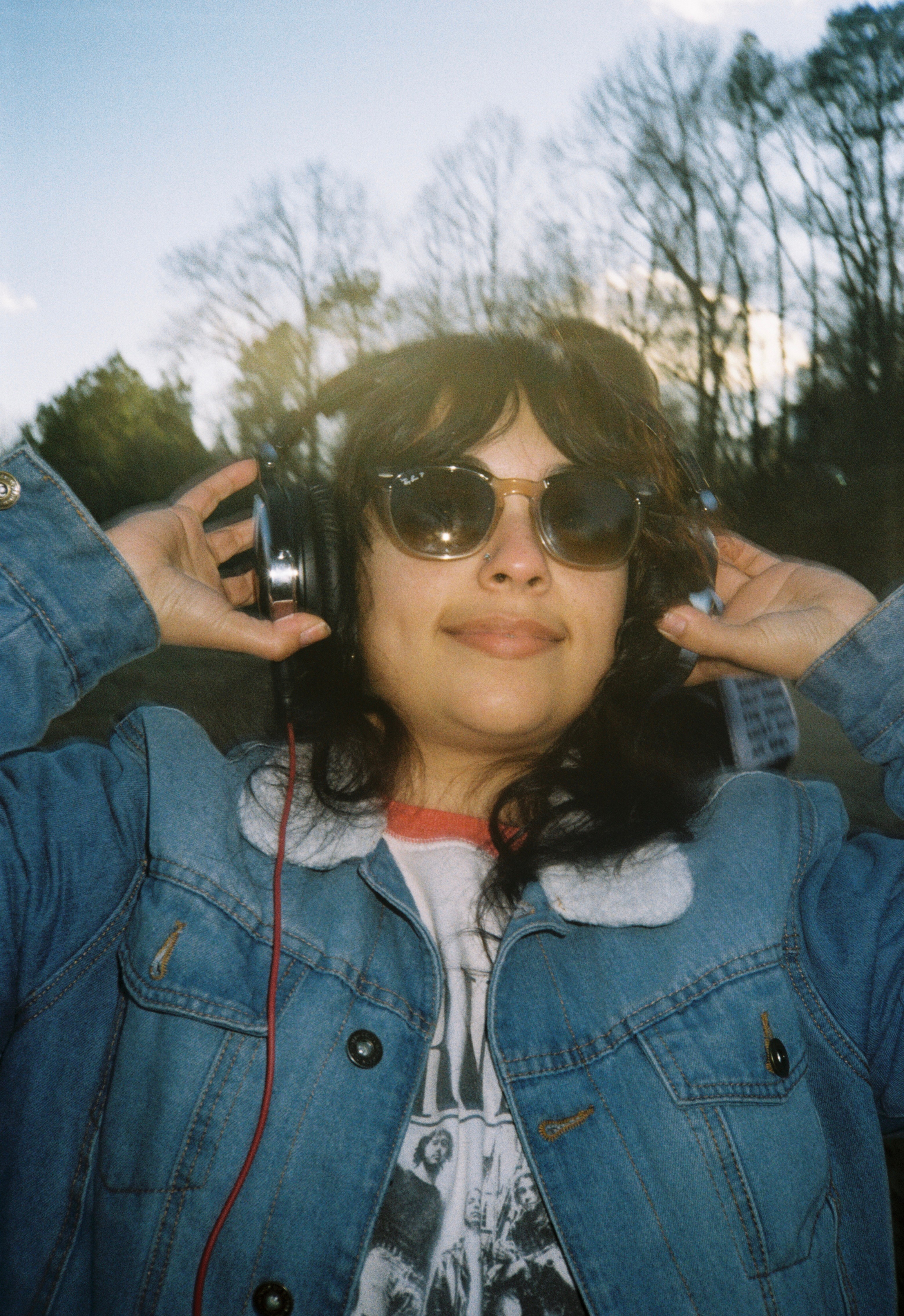 Person wearing sunglasses and headphones outdoors, smiling, with trees and sky in background.