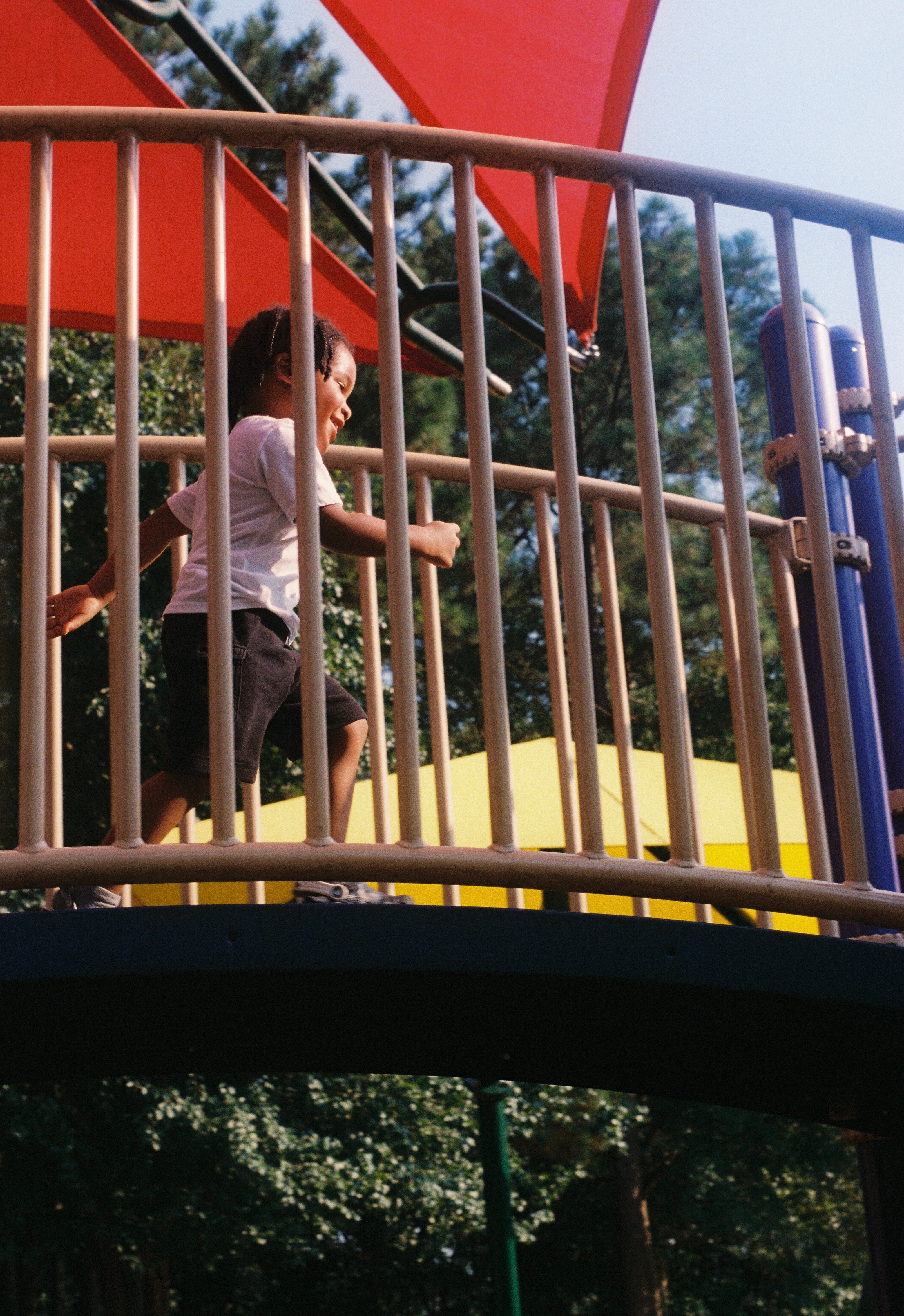 Child running on a playground bridge with metal railings, trees and playground equipment in background.