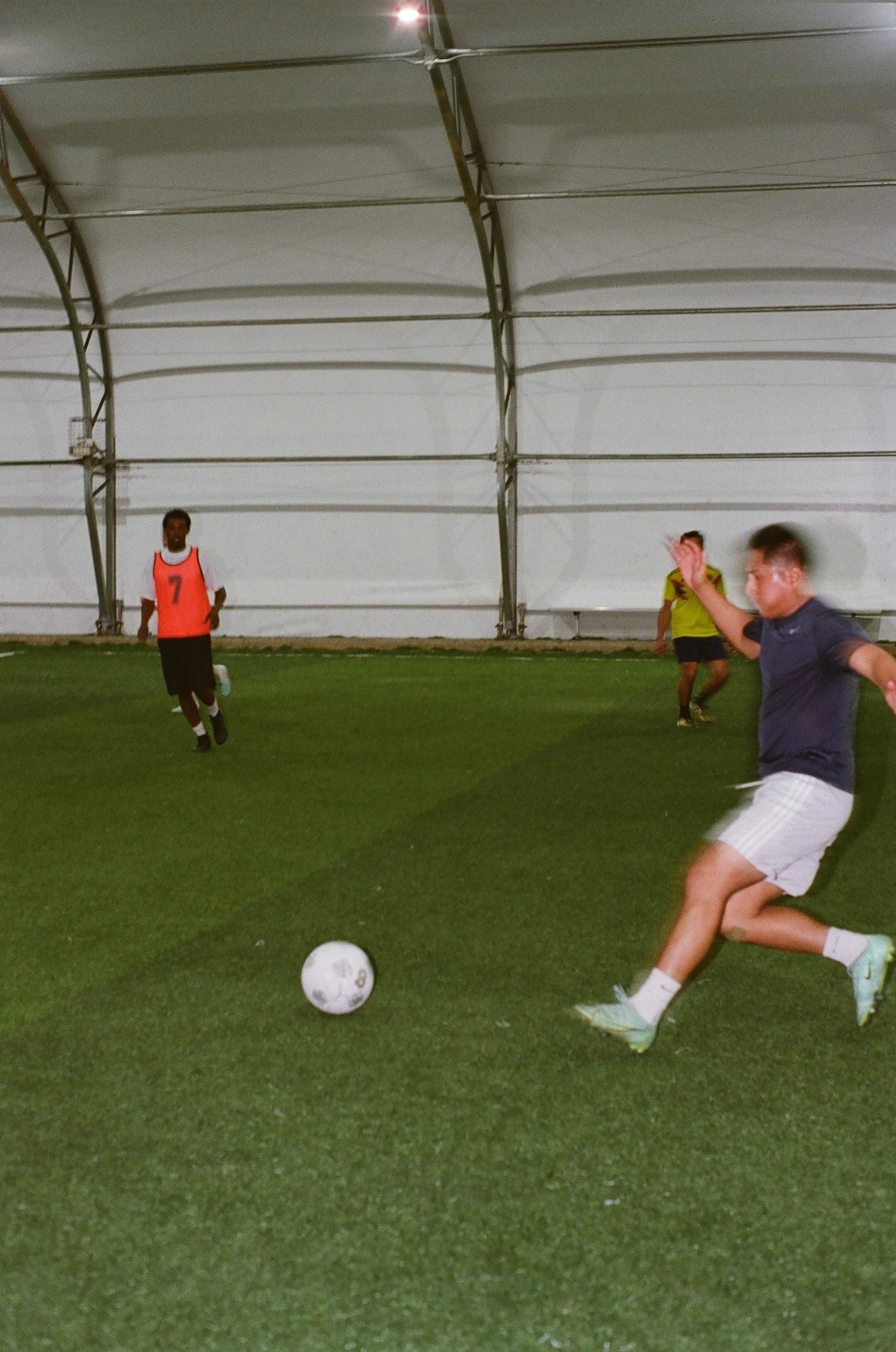 Children playing soccer inside a large indoor facility with a curved ceiling, one child kicking a ball, others in the background.