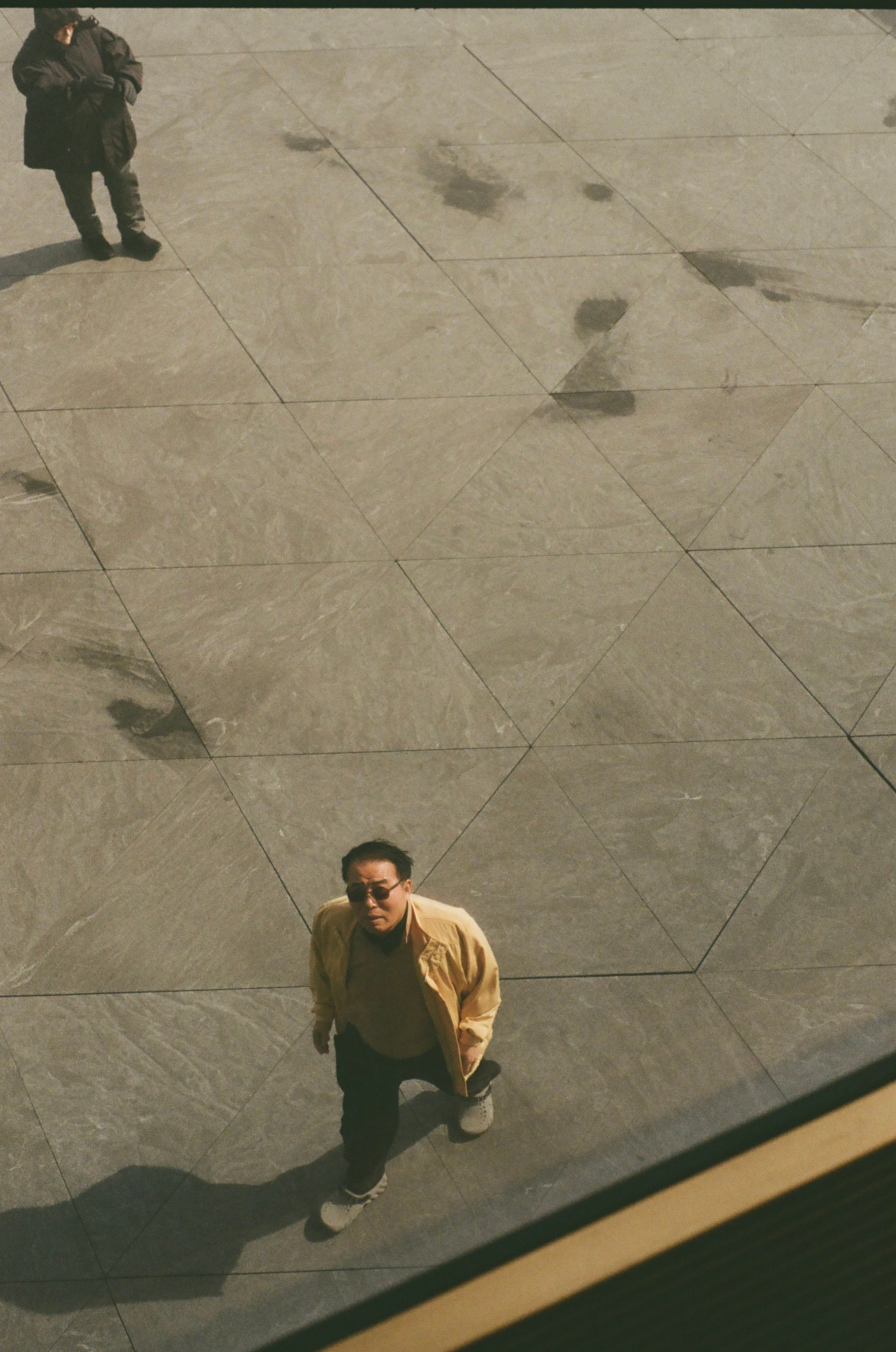 Person standing on a large tiled outdoor surface, looking up at the camera, with a shadow cast on the ground.
