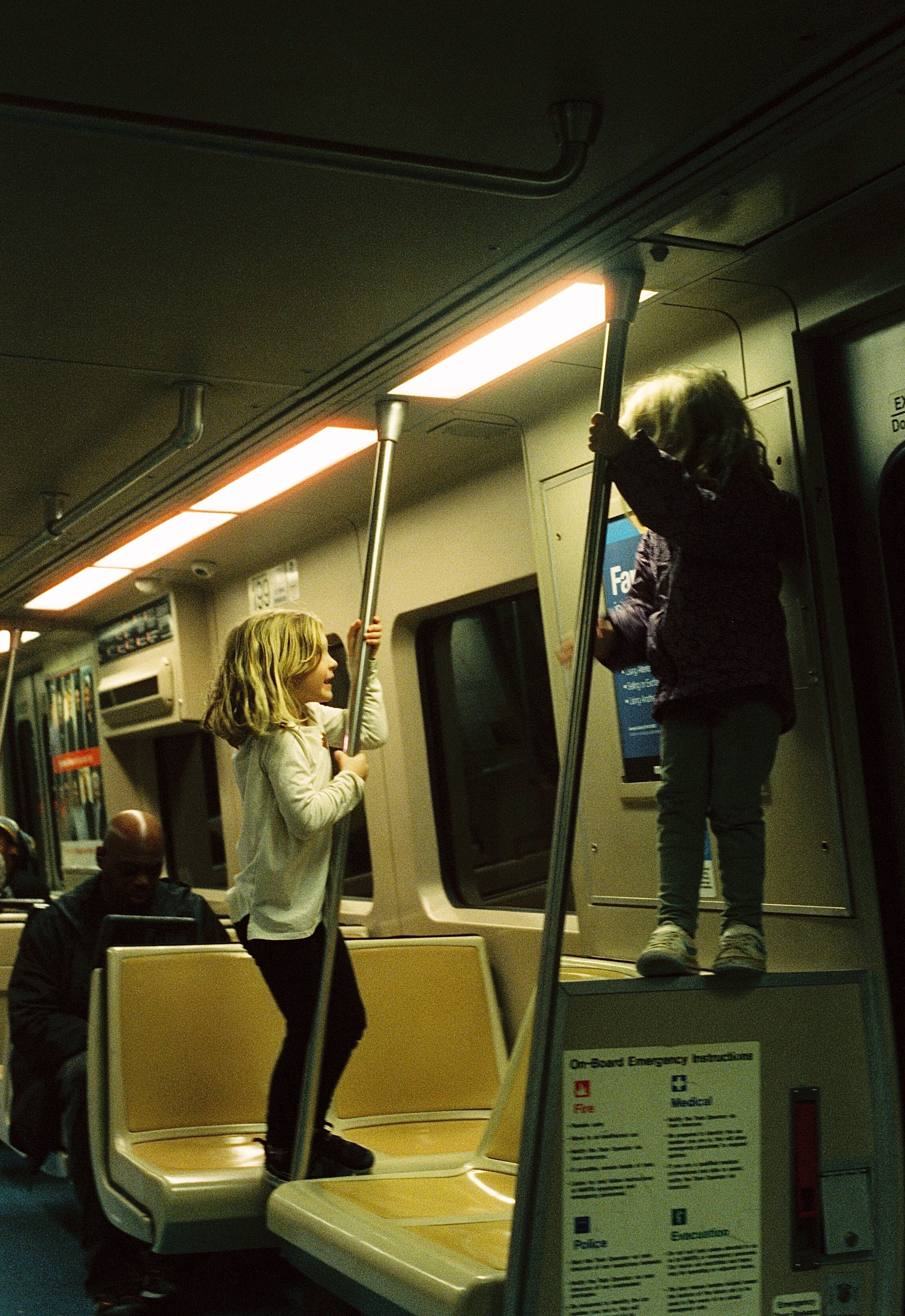 Two children standing inside a subway train, one on a seat and the other holding a pole, with a passenger in the background.