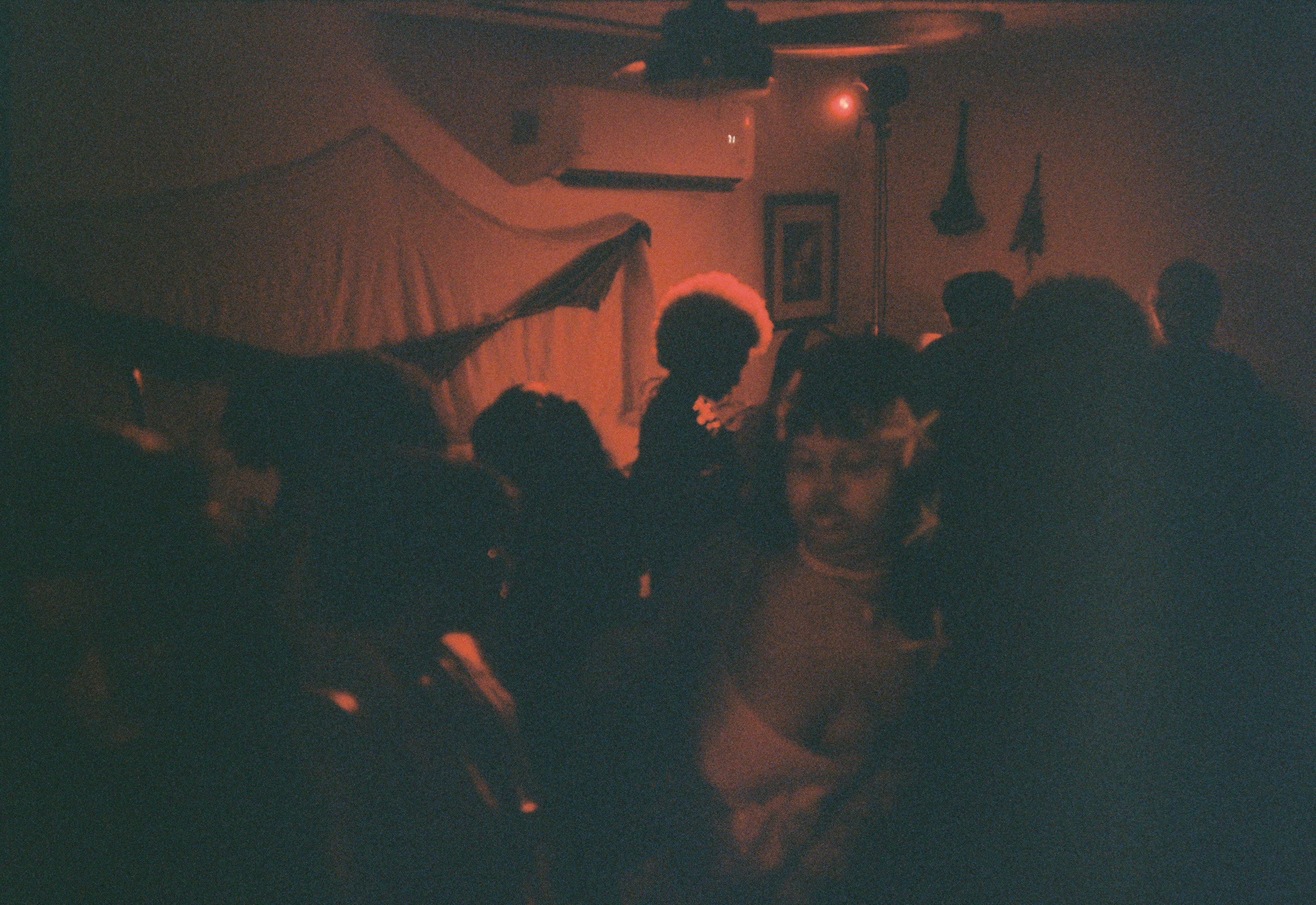 People gathered in a dimly lit room with a draped wall and flags, some faces visible, others obscured by darkness.