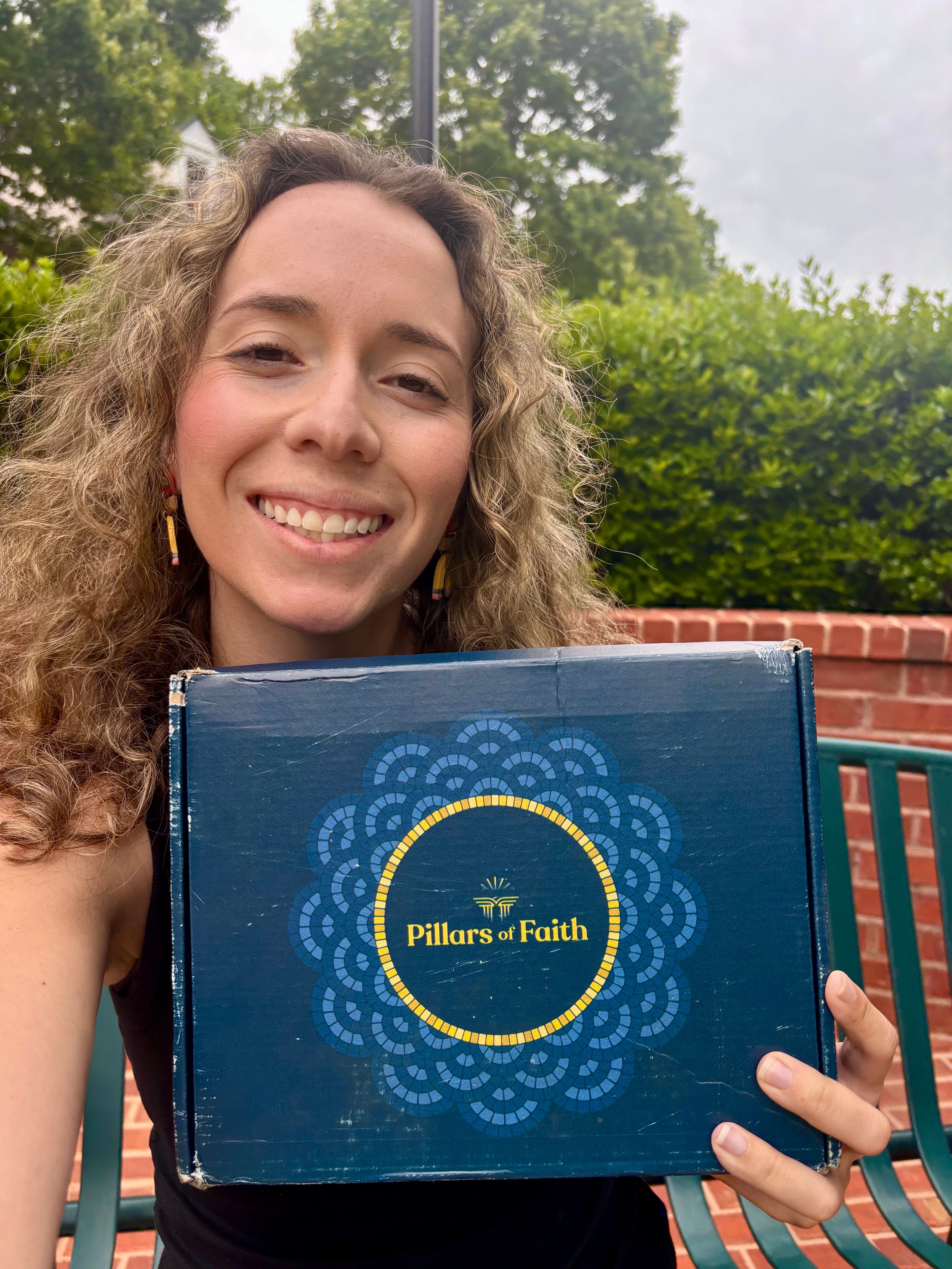 Smiling woman with curly hair holding a blue box labeled 'Pillars of Faith' outdoors, with trees and a brick wall in background.