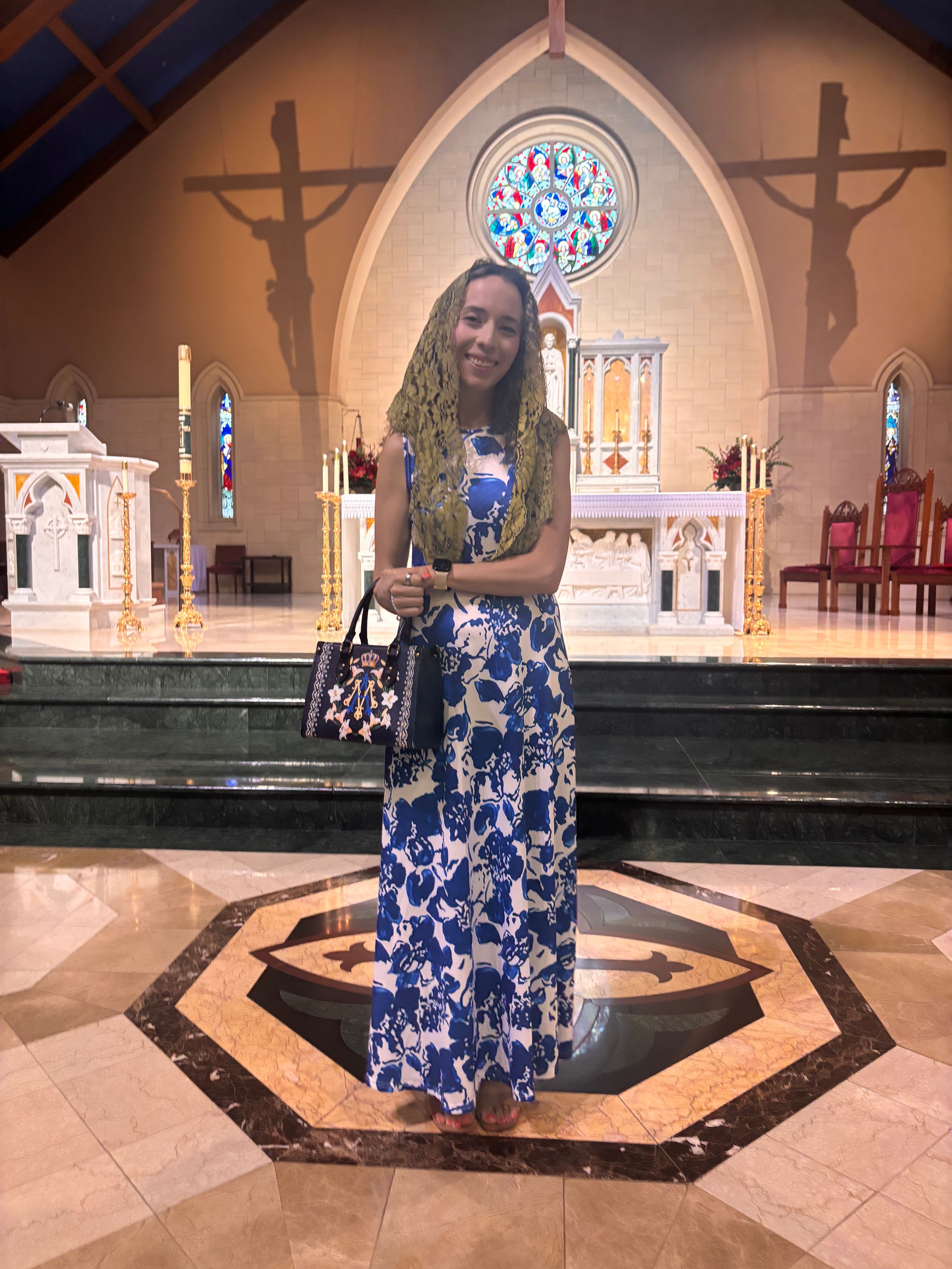 Woman in a blue and white floral dress standing inside a church with stained glass window and altar behind her.