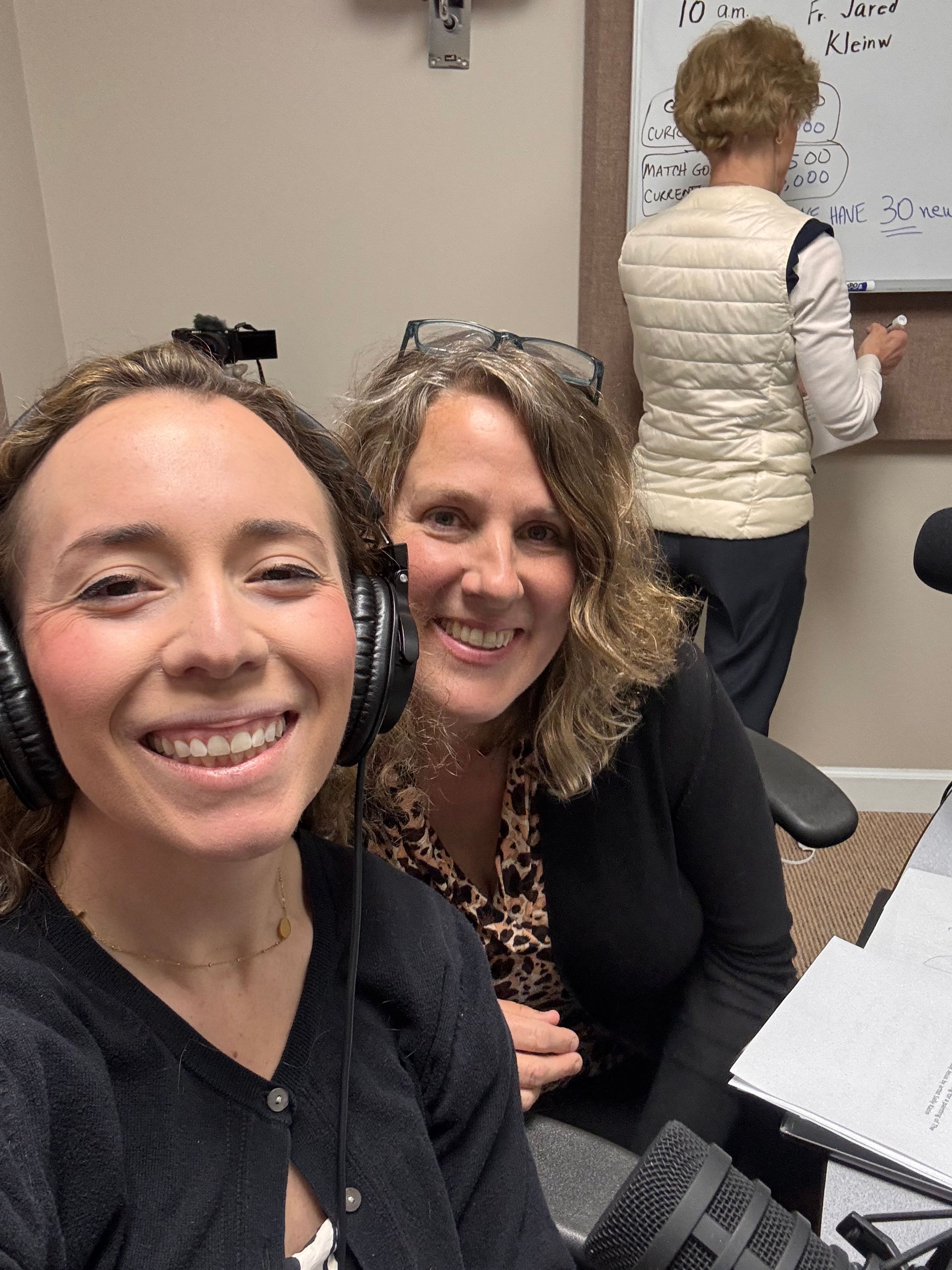 Two women smiling in an office, one wearing a black jacket and the other with curly hair, with a woman writing in the background.