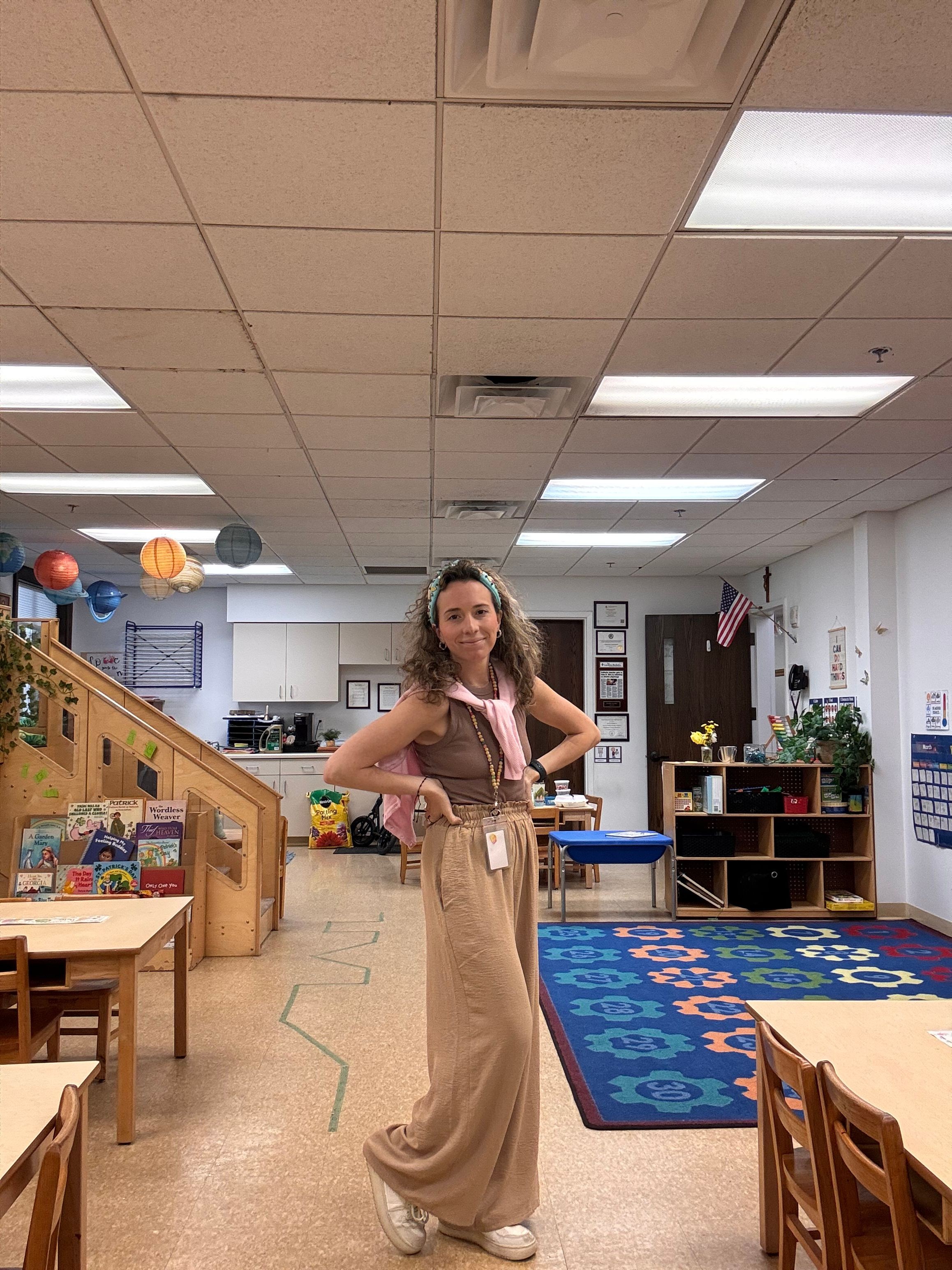 Woman in a pink top and beige pants sitting on a wooden counter in an office or classroom, with colorful decorations and furniture.
