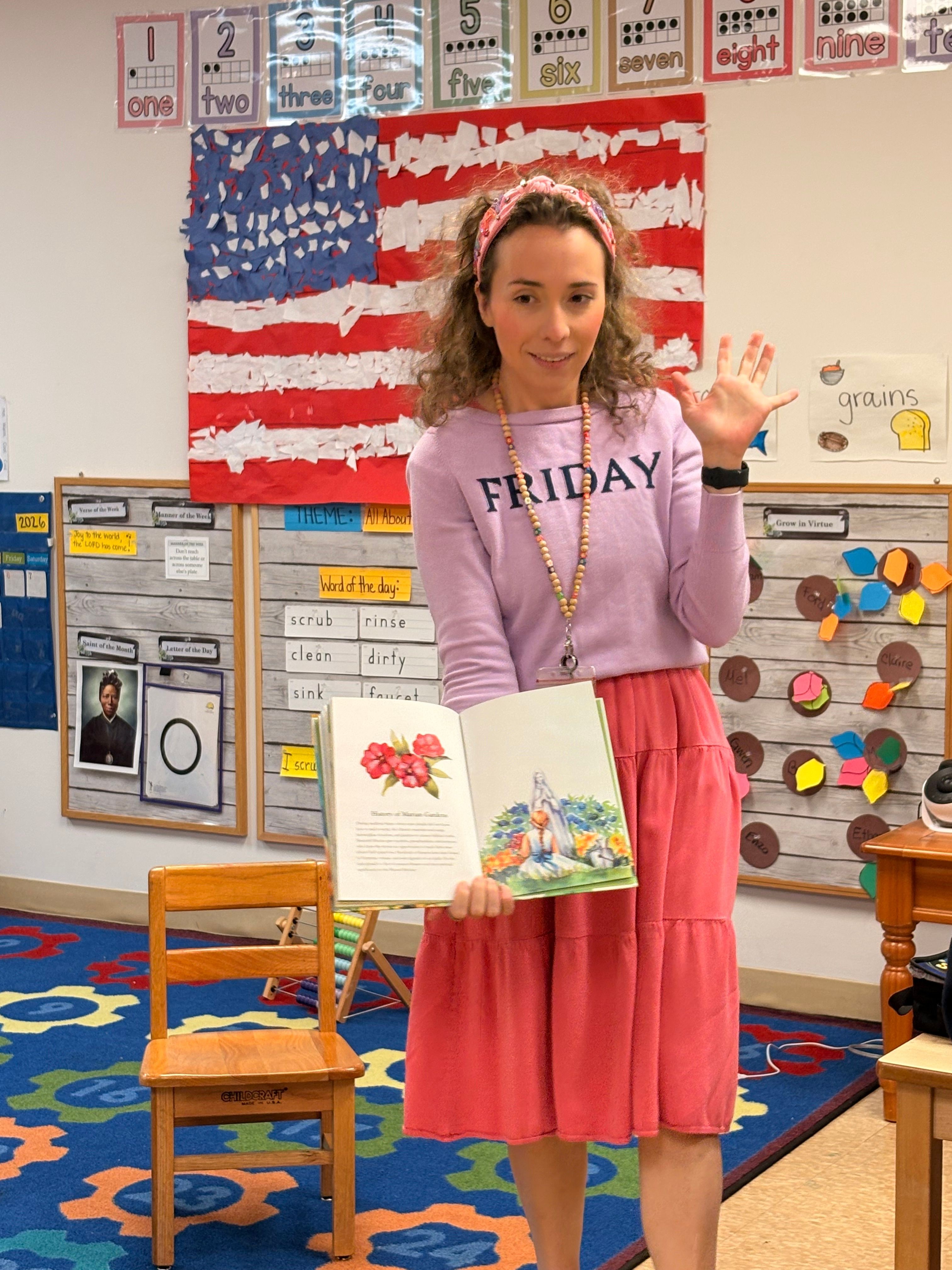 Teacher standing in classroom, holding open book, with American flag display behind, wearing pink skirt and purple top.