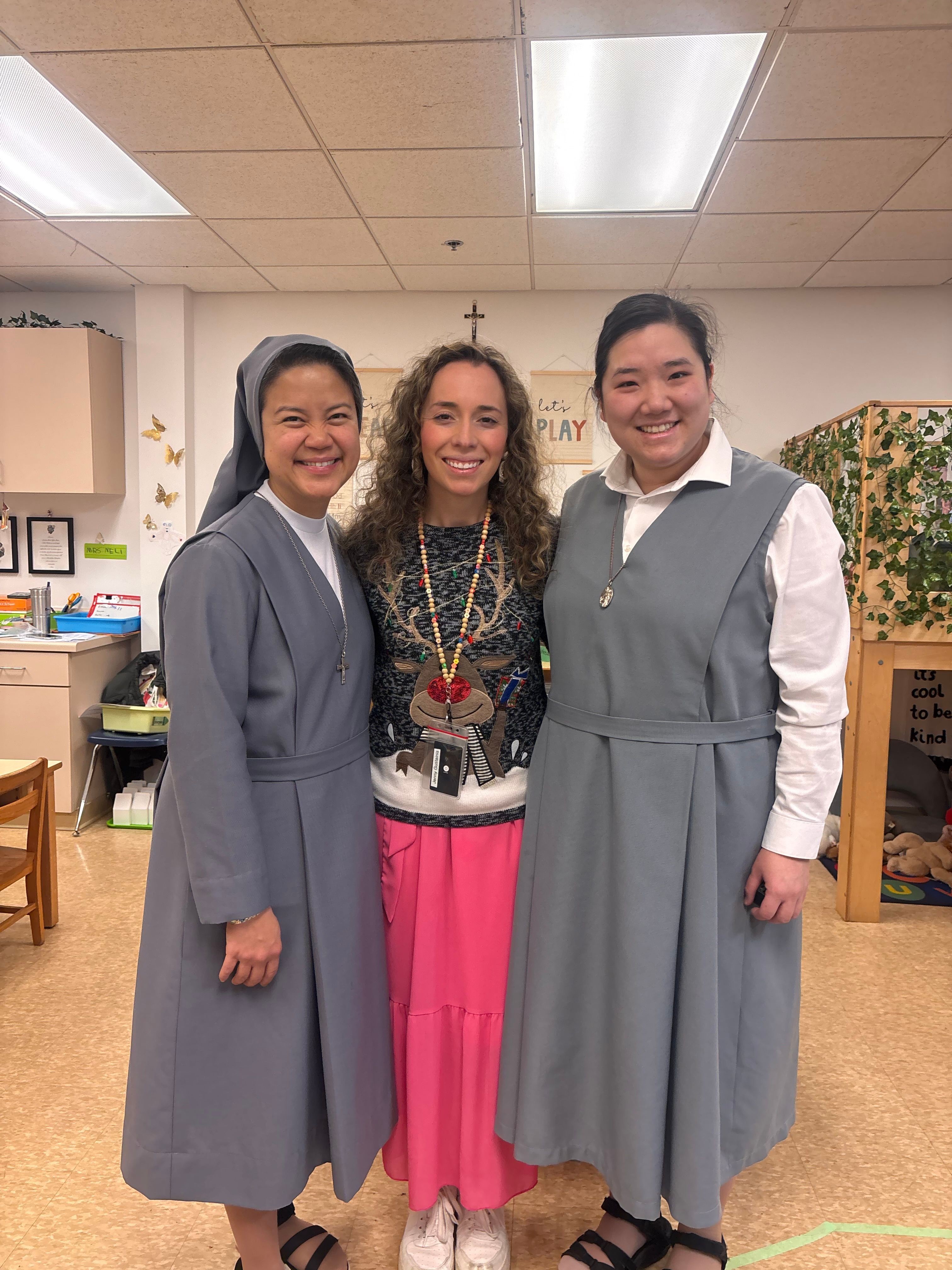 Three women standing indoors, smiling, with a background of office or classroom setting, plants, and wall decorations.