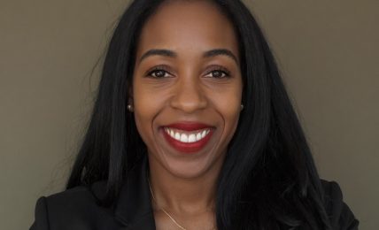 Smiling woman with long black hair, wearing a black blazer and a necklace, against a plain background.