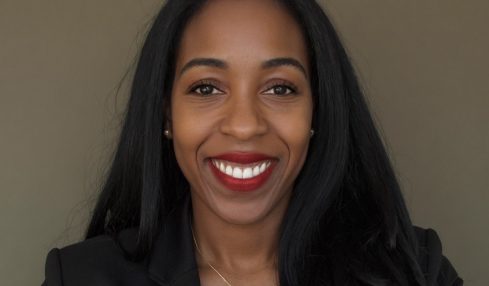 Smiling woman with long black hair, wearing a black blazer and a necklace, against a plain background.