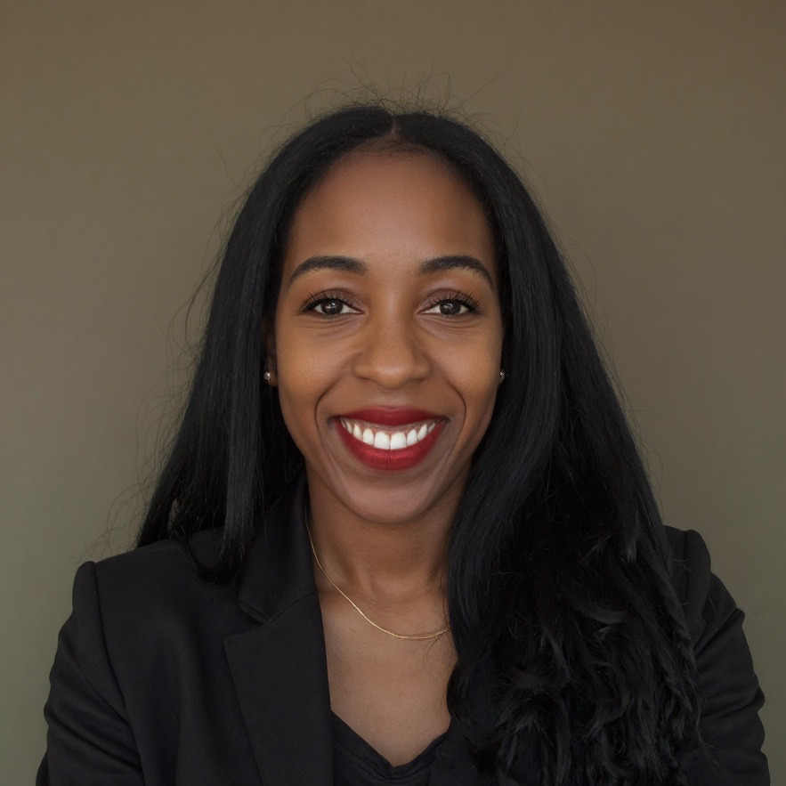Smiling woman with long black hair, wearing a black blazer and a necklace, against a plain background.