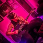 Person playing an electric guitar in a room with pink lighting, wearing glasses and a cap.