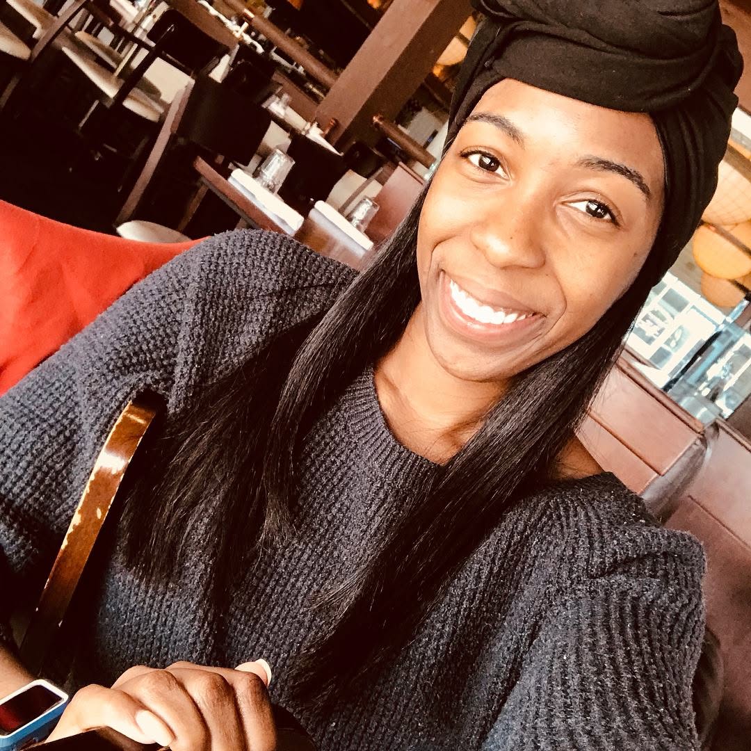 Smiling woman with long black hair wearing a black headwrap and gray sweater, sitting in a restaurant.