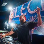 DJ with beard and glasses raises one arm in front of a colorful LED screen at a concert, bright lights shining.