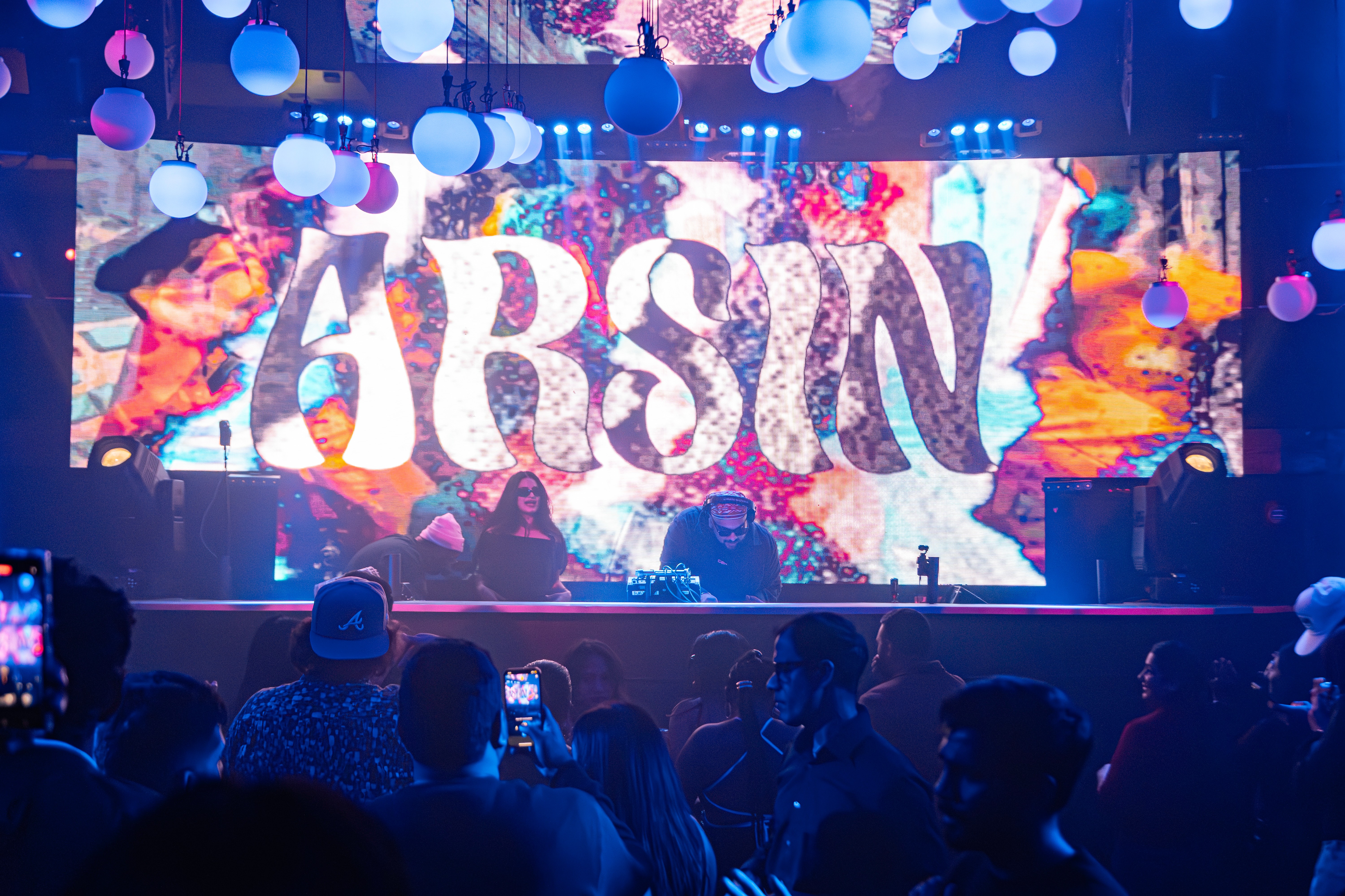 A DJ performs on stage with a colorful background and audience in front, illuminated by blue and purple lights.