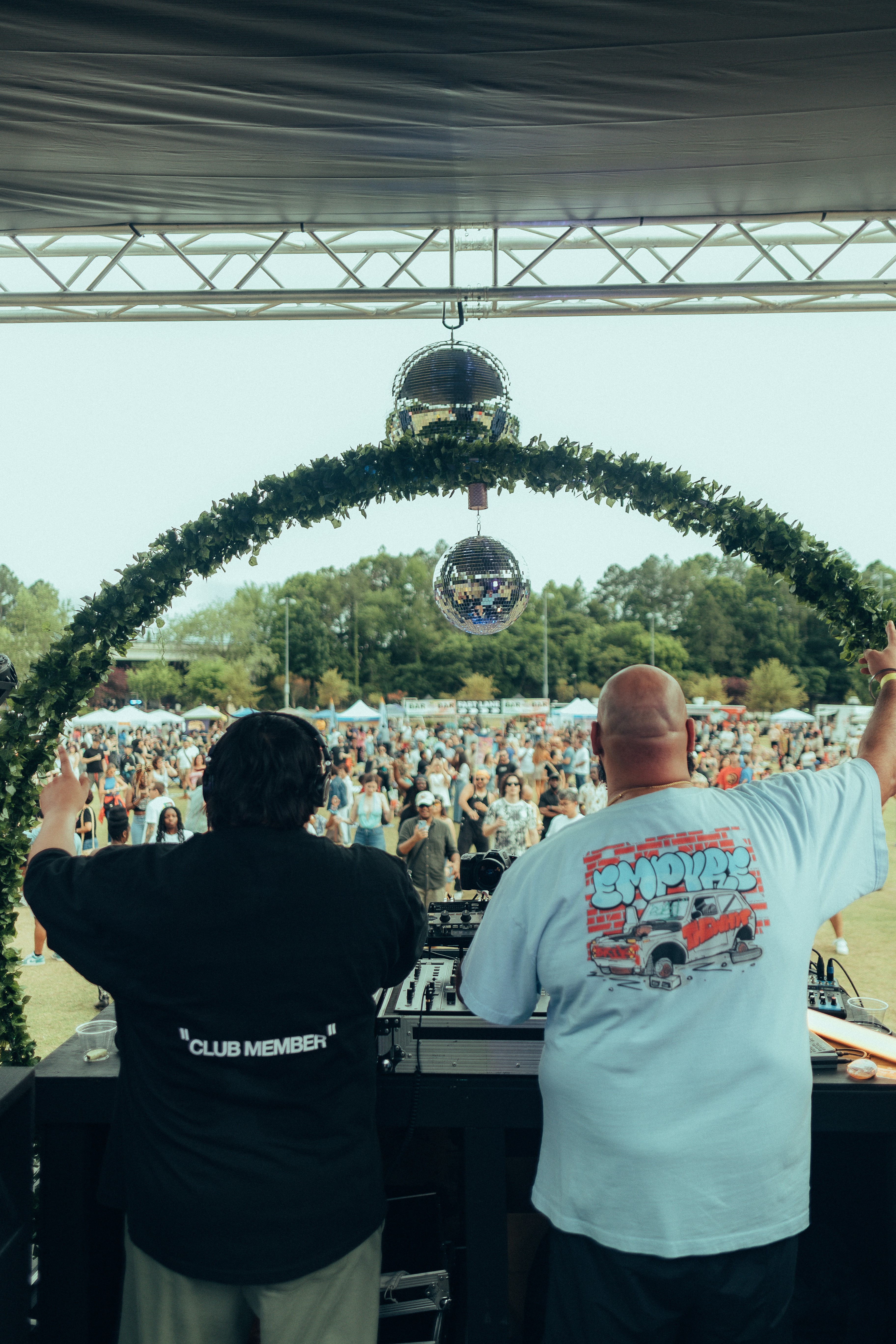 Two DJs perform on stage with a large crowd and decorative arch in the background.