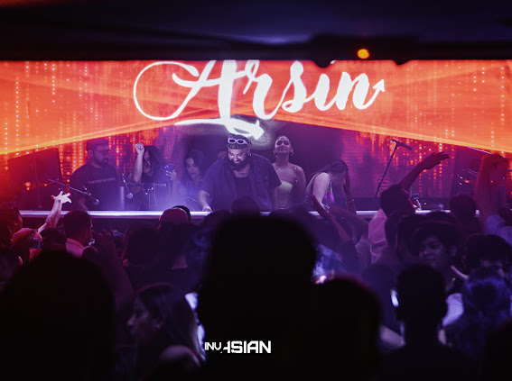 People dancing in a nightclub with a large screen displaying the name 'Hrsin' in bright pink and red lights.