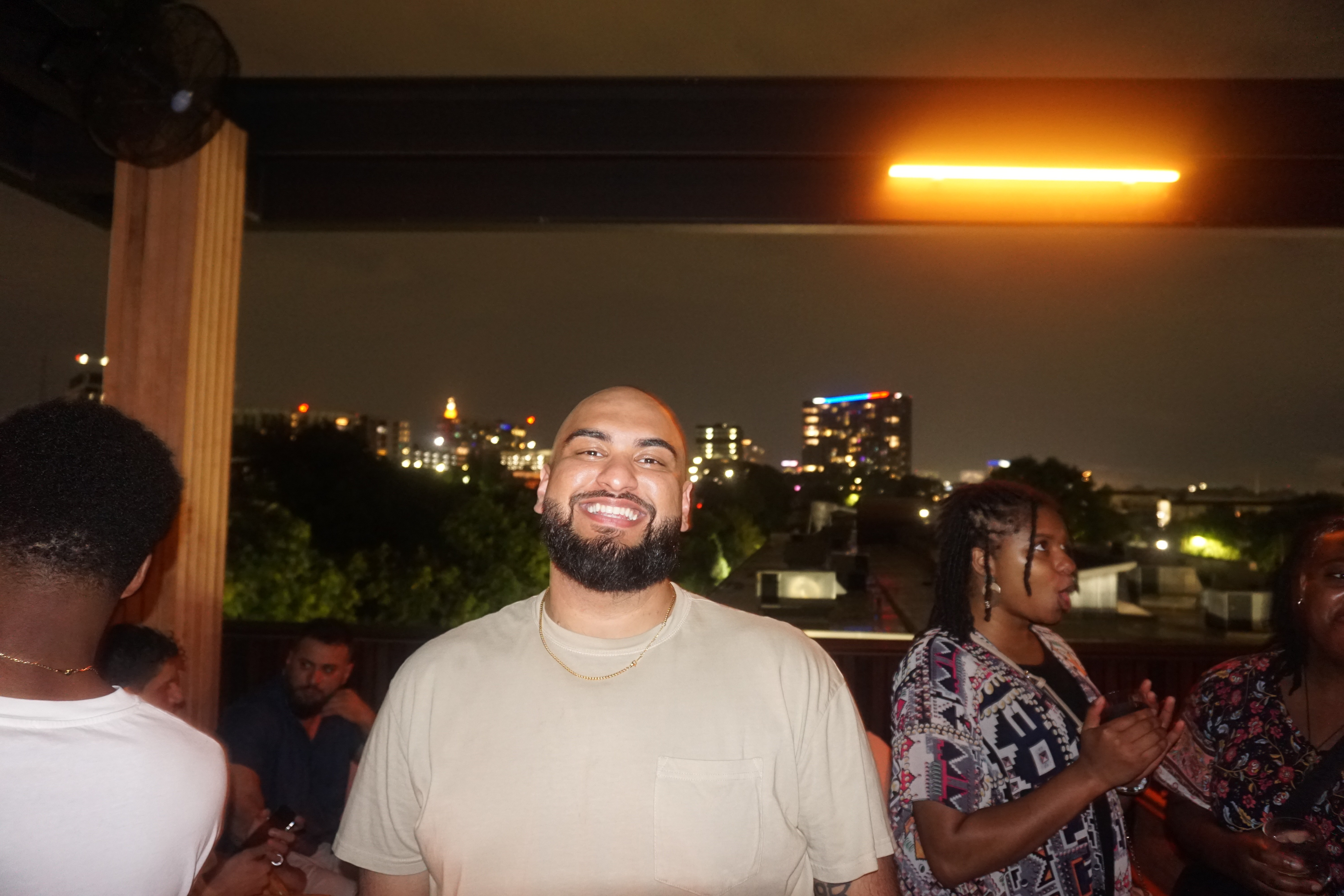 Man with a beard smiling at night with city lights in the background, under a yellow light fixture.
