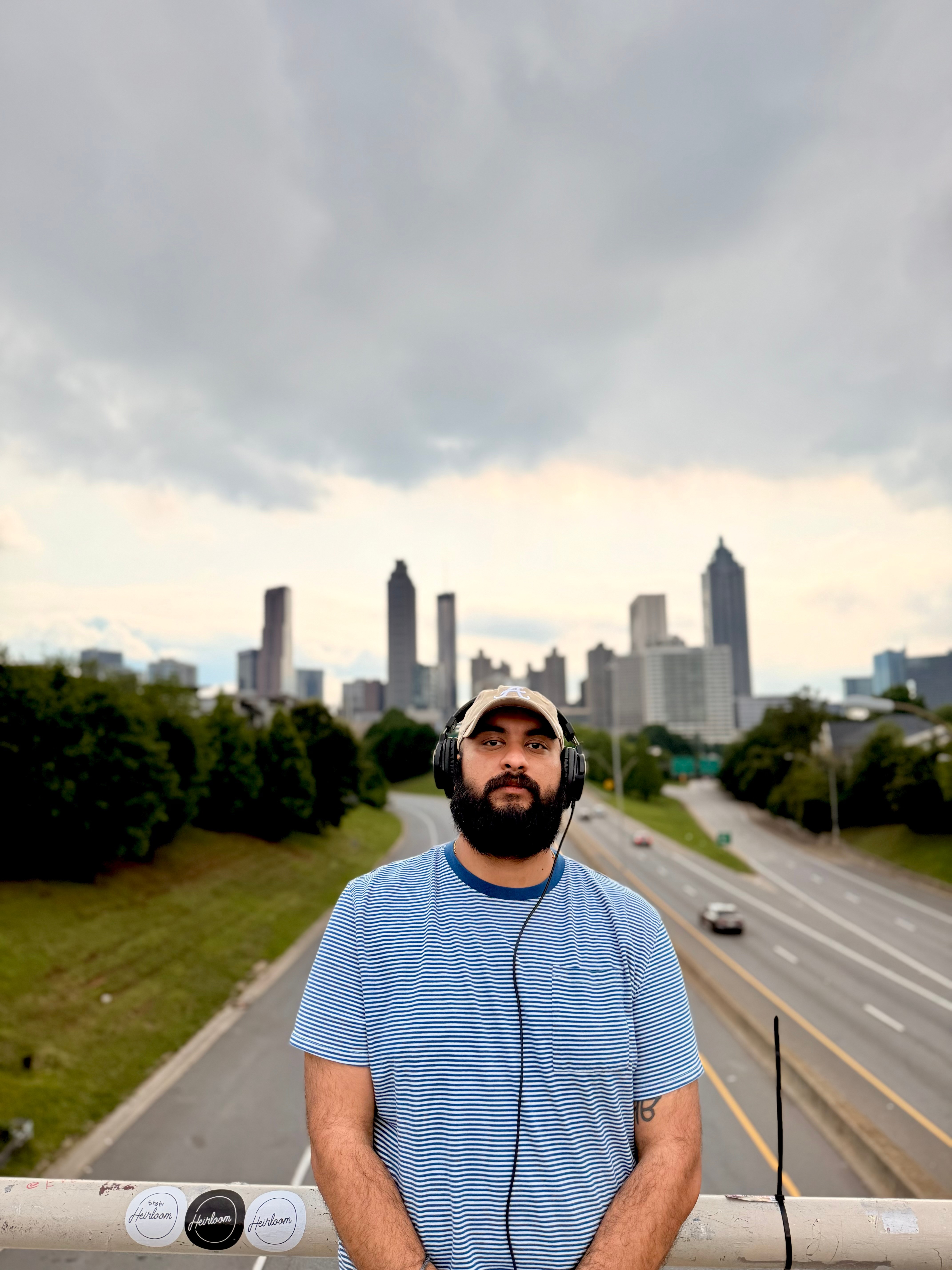 Young man with a beard wearing a blue shirt and white cap, standing outdoors with city skyline and cloudy sky behind him.