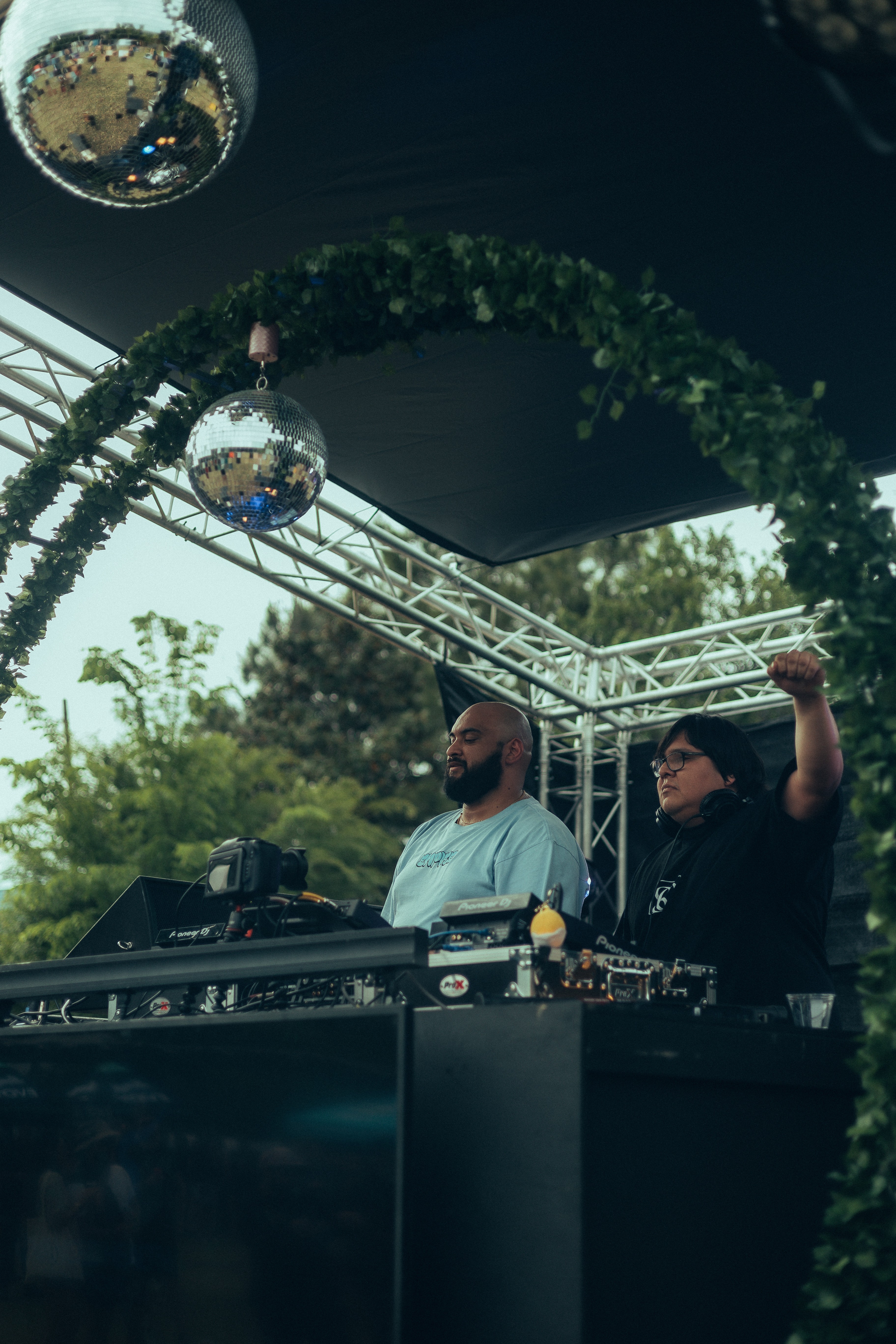Two DJs perform under a decorated arch with greenery and hanging ornaments outdoors, with trees in the background.